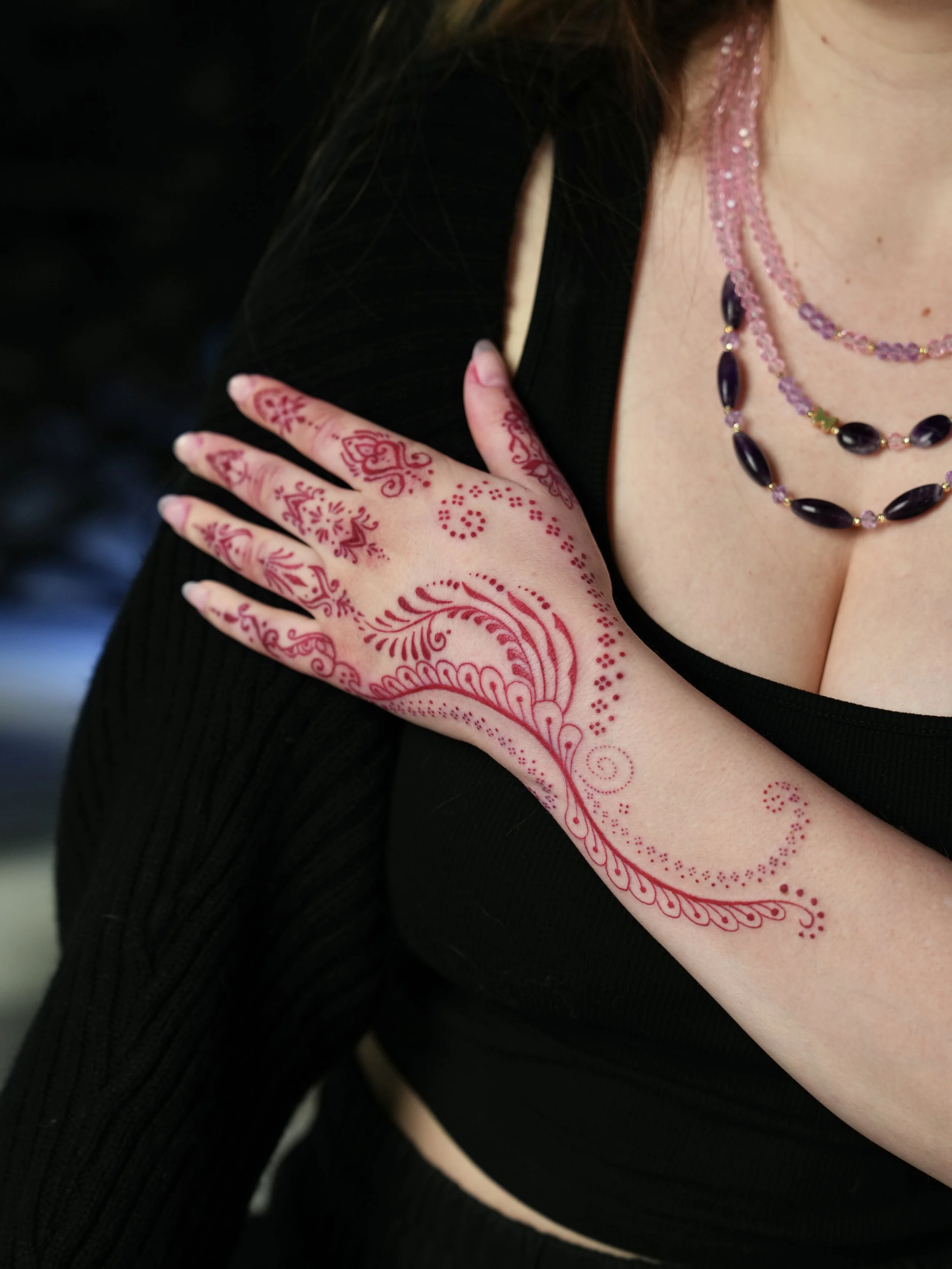 A woman with henna decorated hand on her shoulder, wearing layered beaded necklaces and a black outfit.