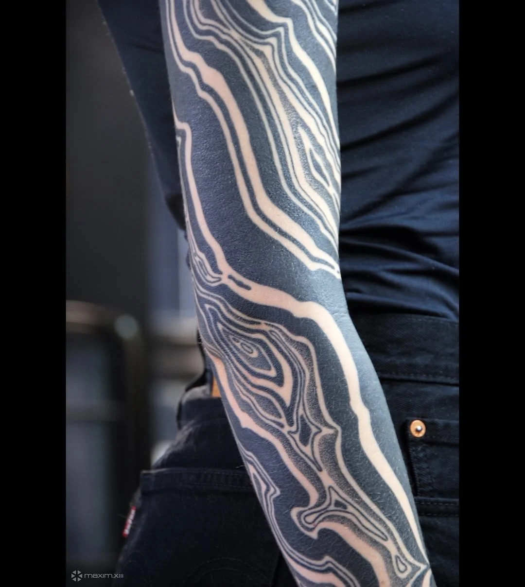 Close-up of a person's arm with a black and white abstract patterned tattoo, wearing a black shirt and pants.