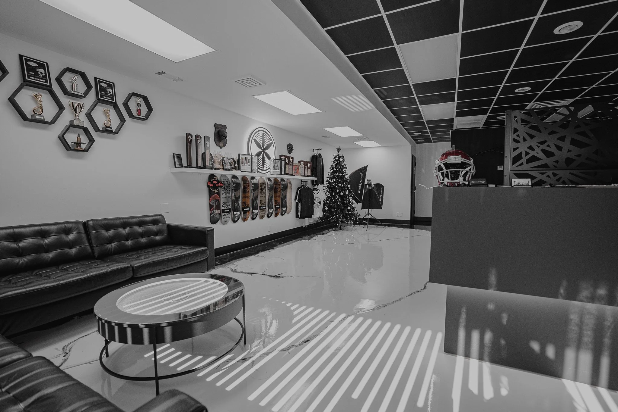 Indoor space with black leather couches, a round coffee table, a decorated Christmas tree, skateboard decks on the wall, trophies and framed awards, photography equipment, and sports memorabilia, with stark lighting and shadow patterns on the floor.