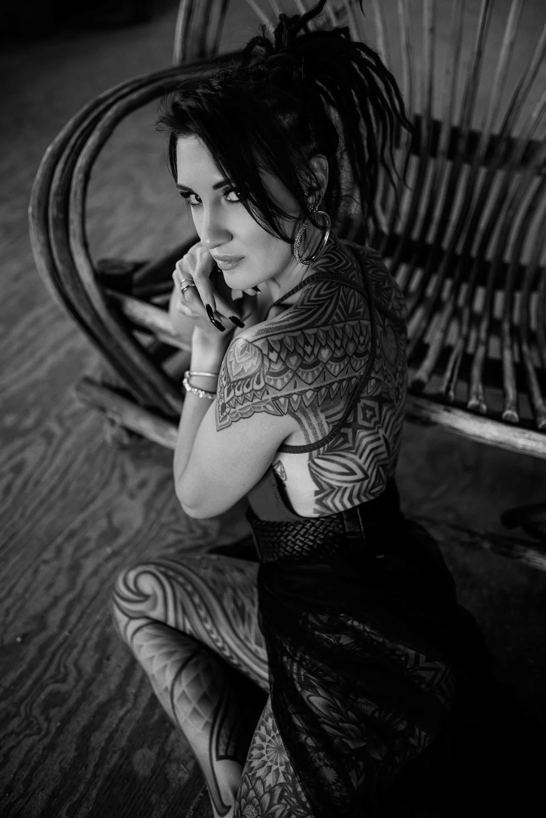 A woman with dreadlocks and tattoos poses on a wooden floor next to a wooden lounge chair, looking up at the camera.