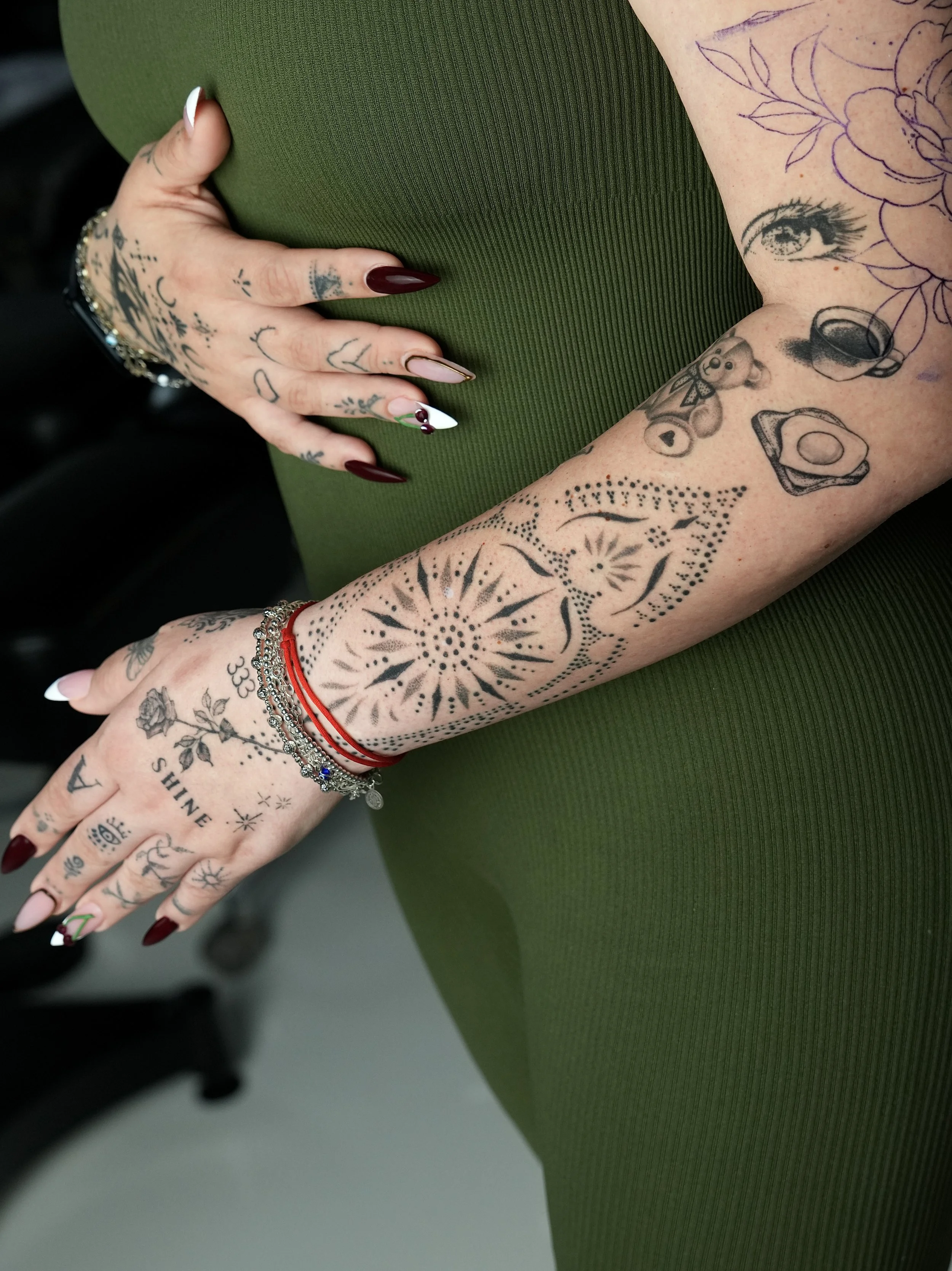 Close-up of a person's arm and hand, showing various tattoos including flowers, a teddy bear, an eye, and abstract designs, with jewelry and a green textured dress.