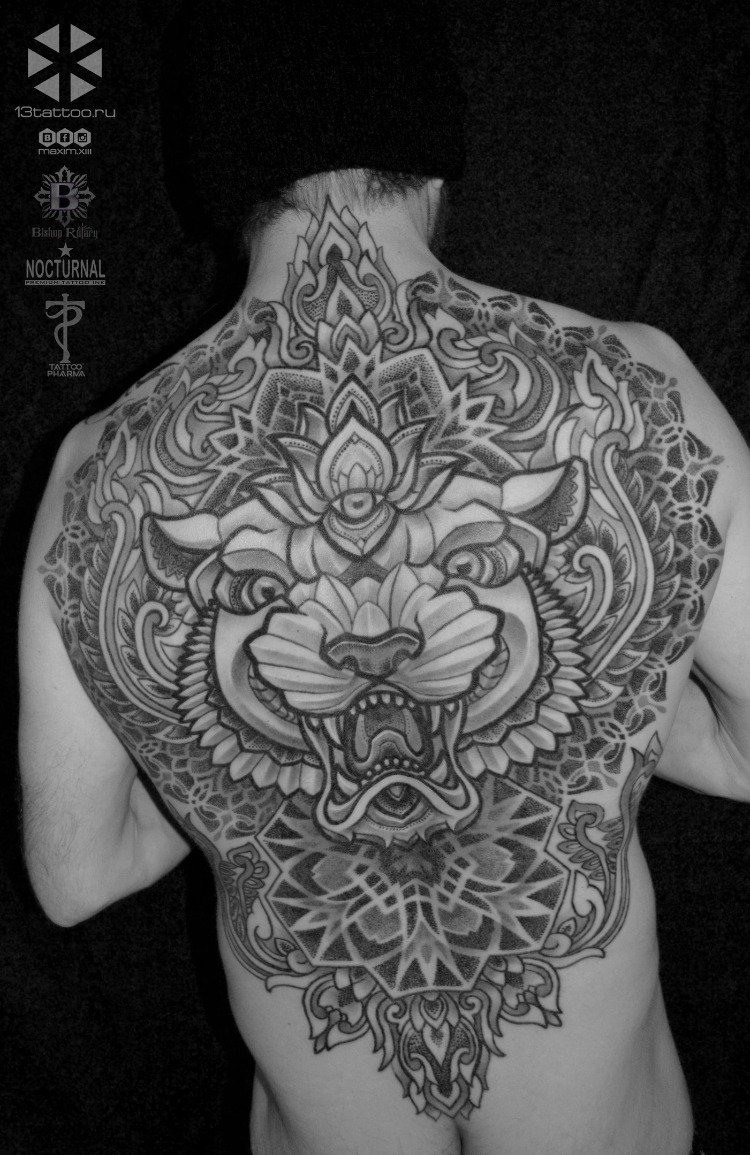 Black-and-white photo of a man's back with a large, intricate tattoo of a fierce tiger's face with a third eye on its forehead, surrounded by ornamental patterns and designs.