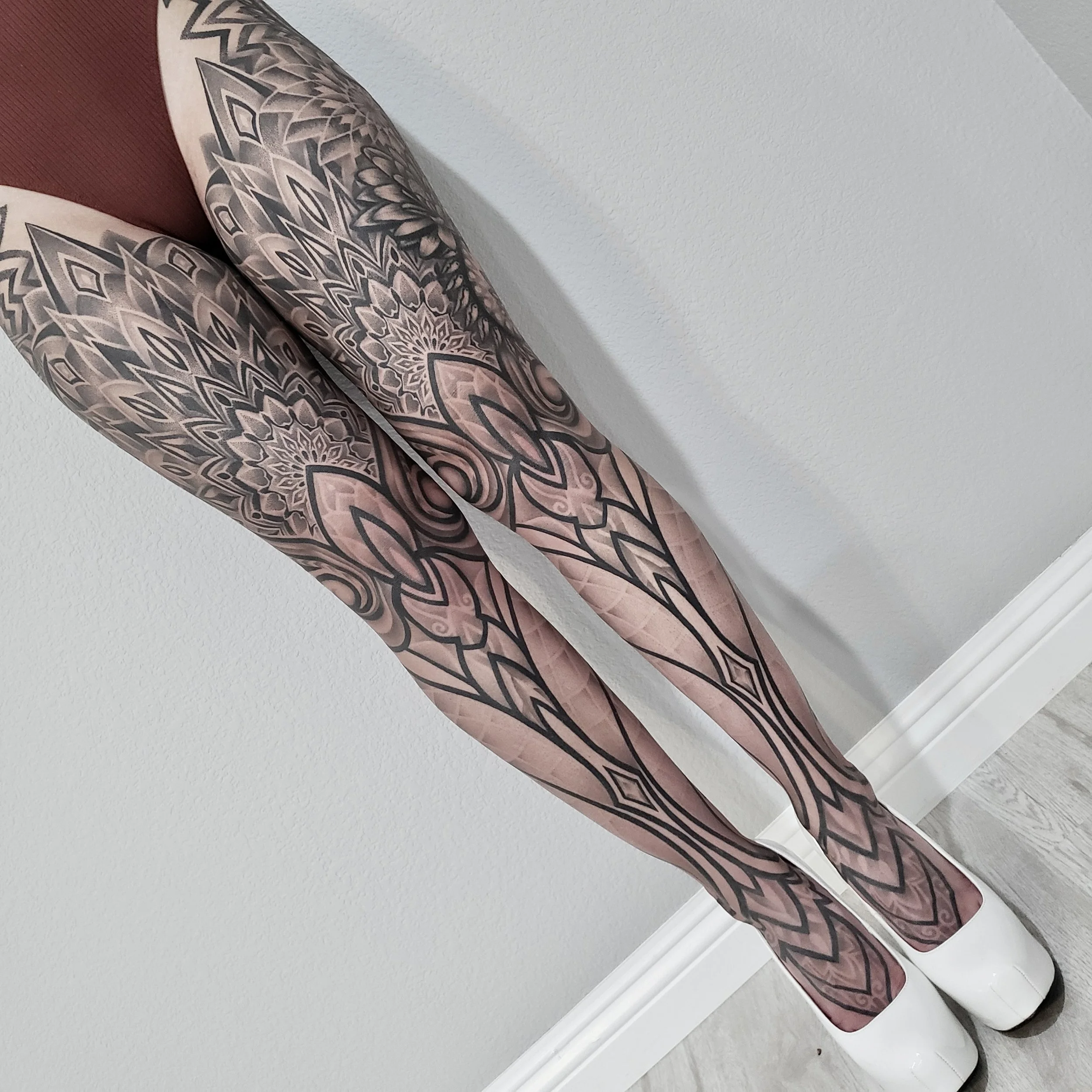Close-up of tattooed legs and feet wearing white high heels against a plain white wall.