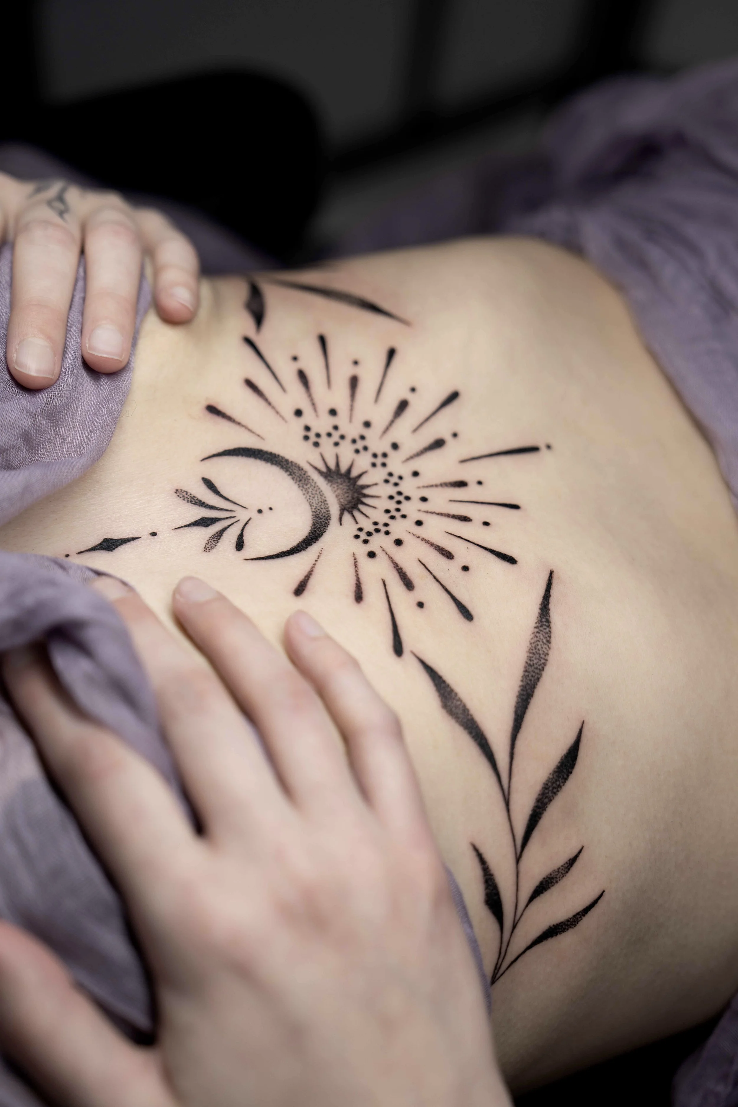 Tattoo of a crescent moon, star, and fireworks on a person's side with hands touching the skin.