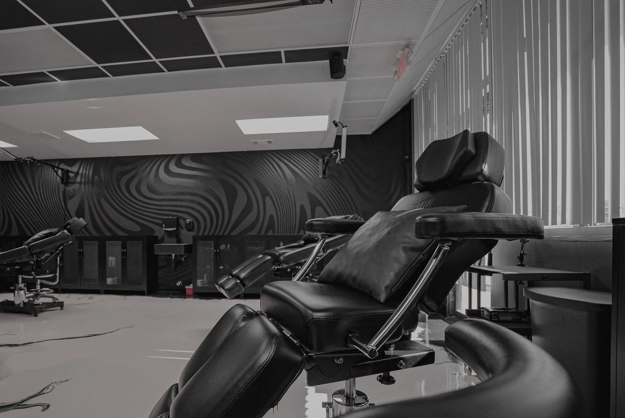 Interior of a clinic or medical treatment room with black medical chairs, including one with a pillow, and a decorative black and white wall in the background.