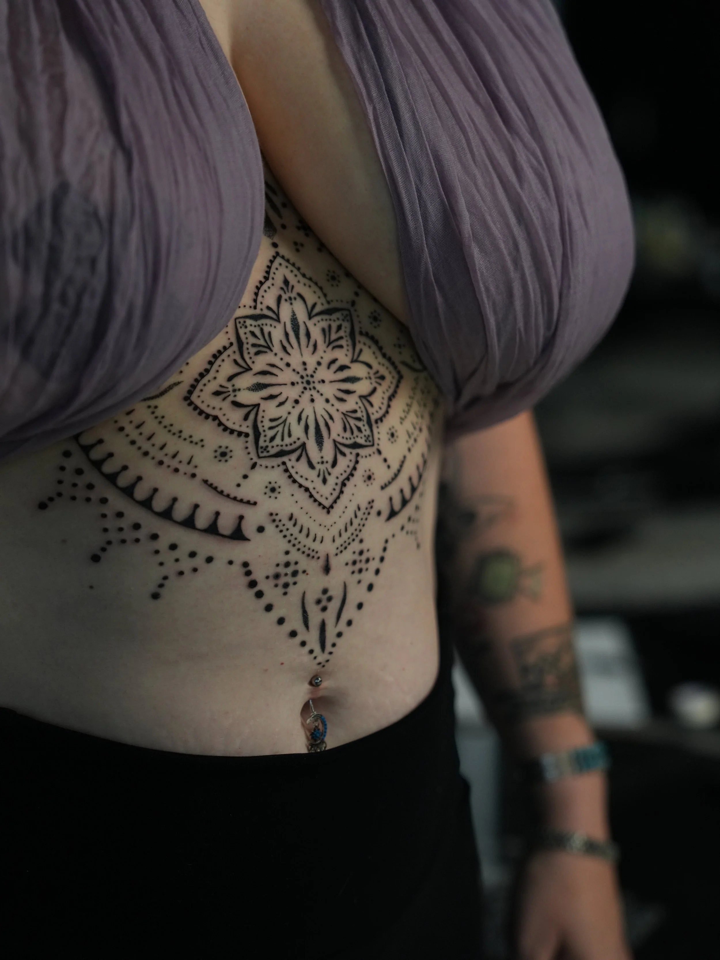 Close-up of a woman's chest with black tattoo design, purple top, navel piercing, and bracelets on her arm.