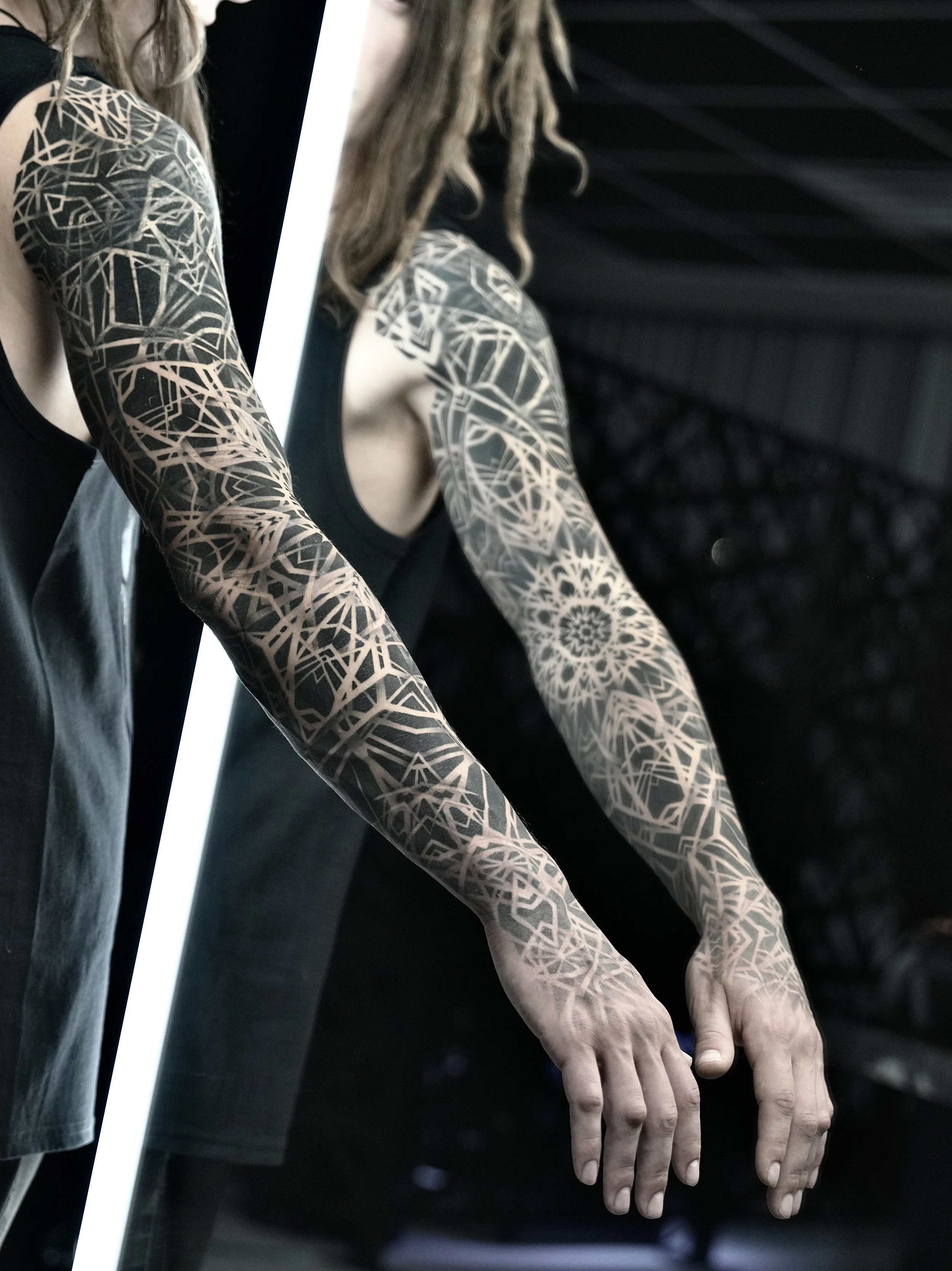 Two people with patterned arm tattoos stretching their arms forward. Both are wearing black sleeveless tops. The background is dark with some structural elements.