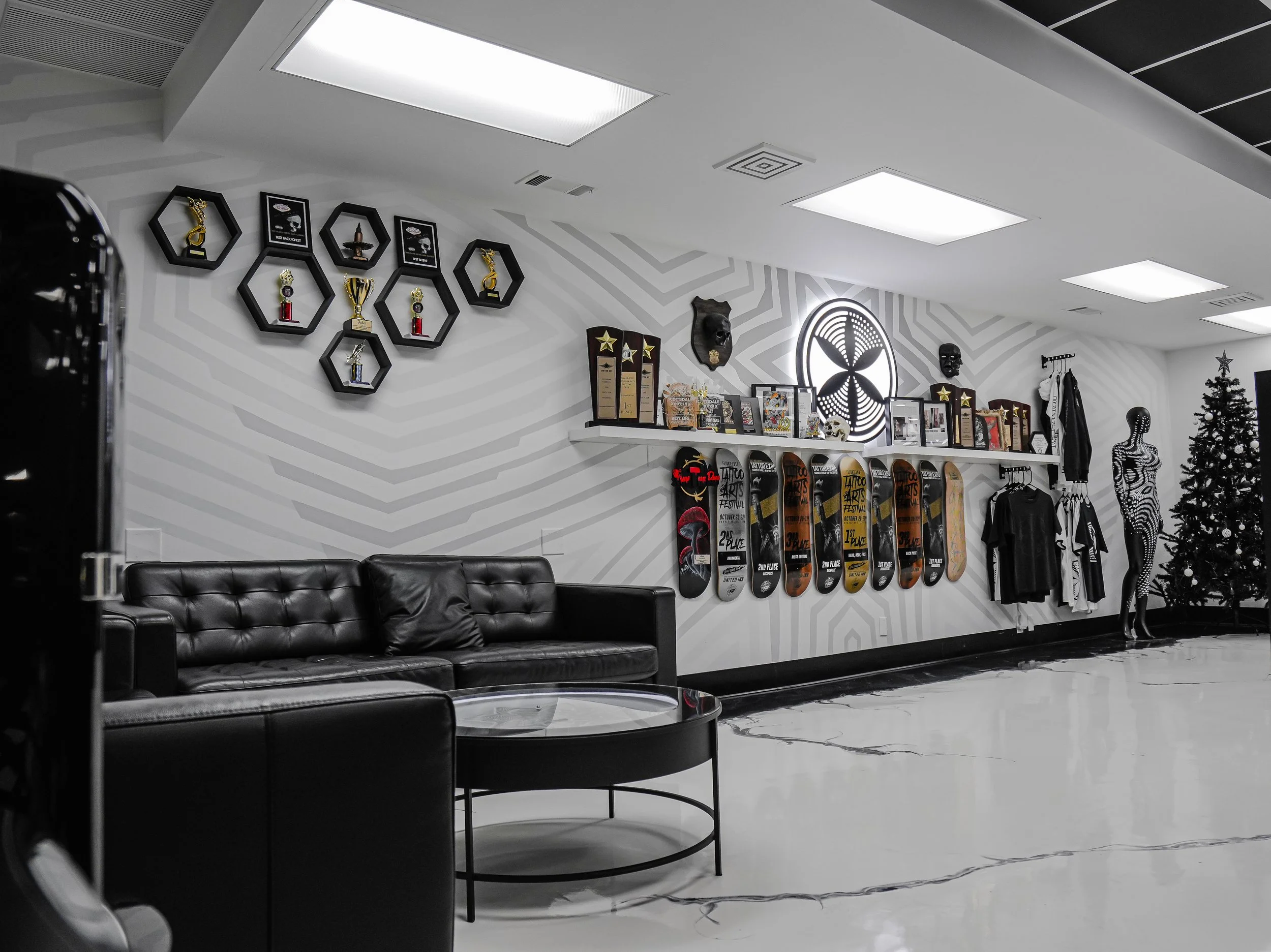 Interior of a modern lounge or reception area with black leather sofa, skateboard awards on the wall, trophies, framed pictures, mannequins, and a decorated Christmas tree.