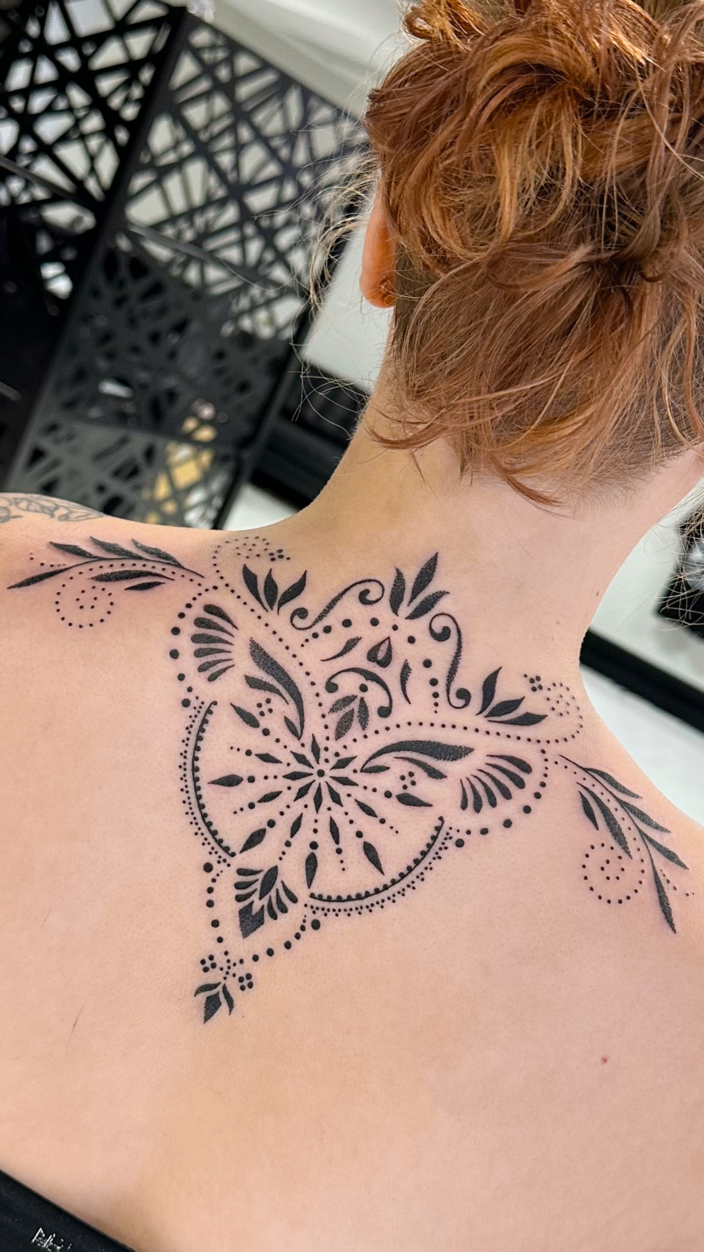 Close-up of a person's upper back and shoulder with a fresh, intricate black tattoo design consisting of floral and geometric patterns.