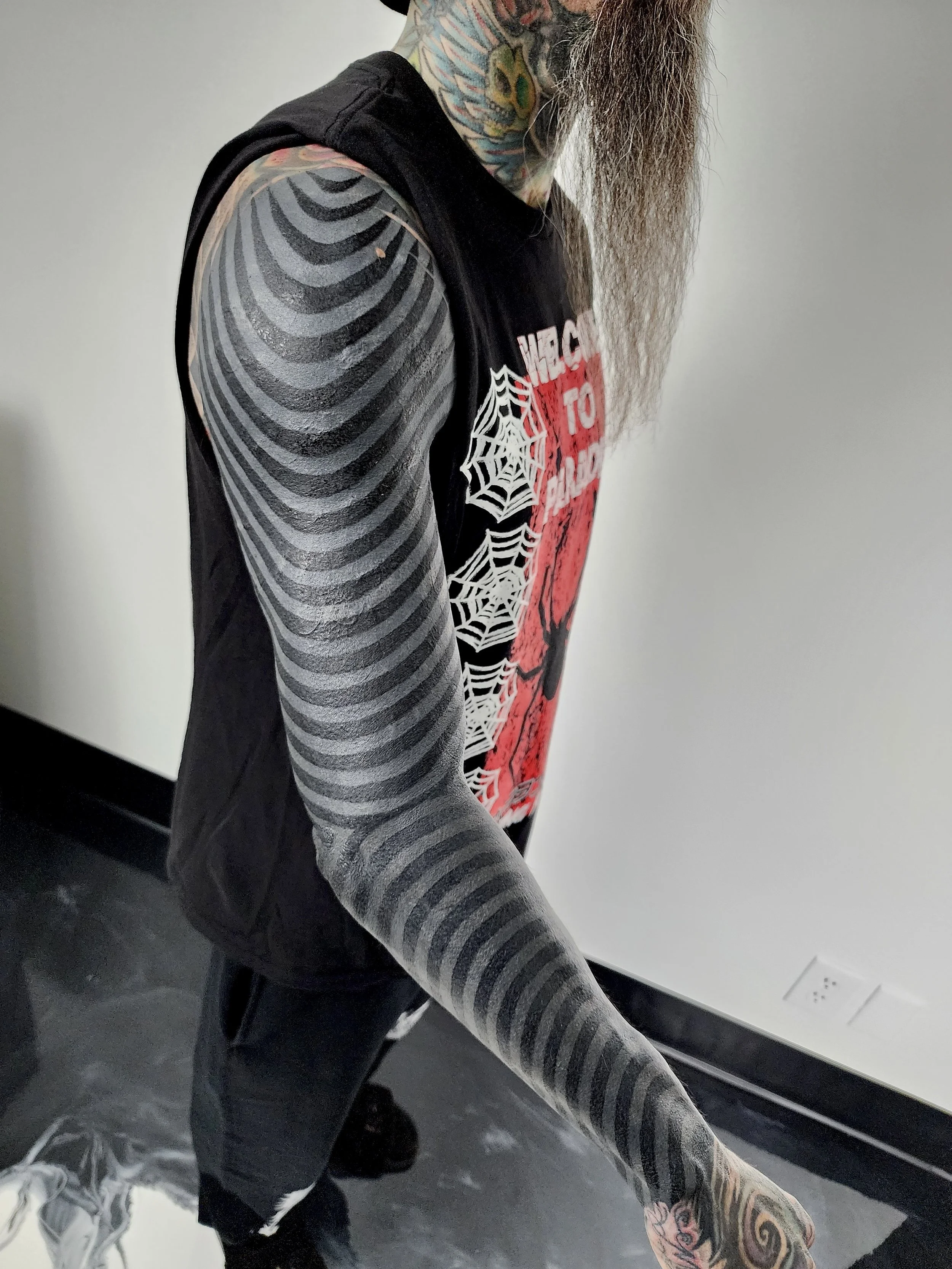 A person with extensive tattoos on their arm and neck, wearing a black sleeveless hoodie with graphic prints, standing against a plain wall.