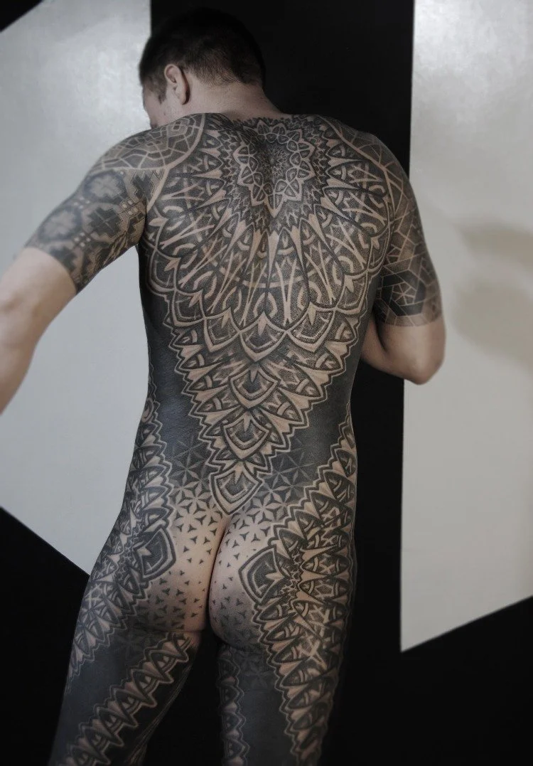 Back view of a man with extensive black and gray geometric tattoos covering his entire back, arms, and legs.