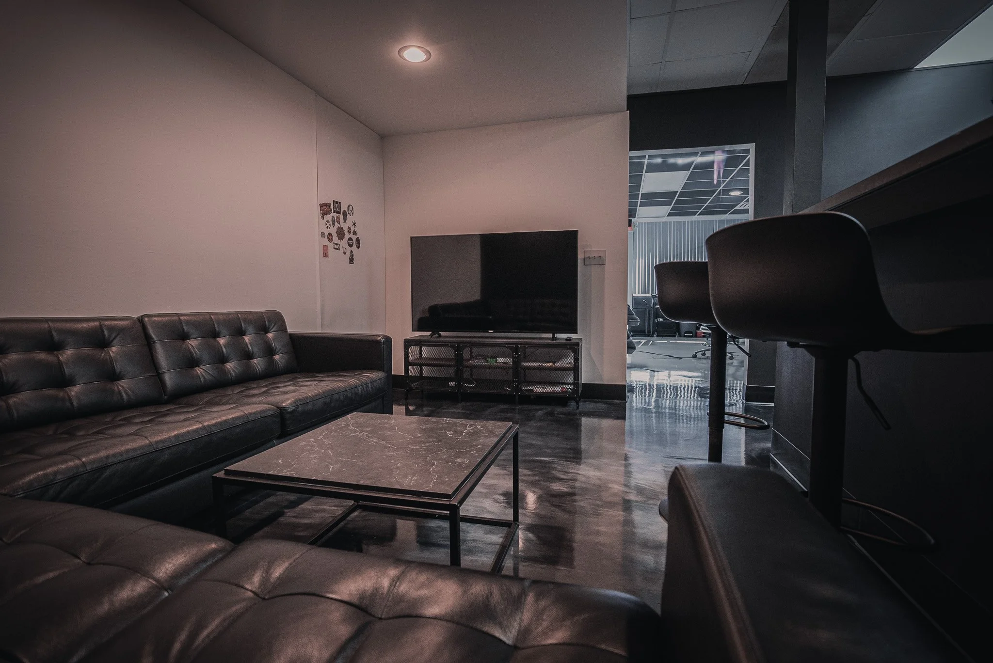 Modern lounge area with black leather sofa, black TV on a stand, marble coffee table, bar stools, and a wall with small decorations, in a setting with polished floor and dim lighting.