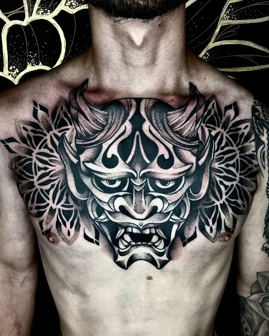 Tattoo of a fierce, stylized face with pointed ears and bared teeth on a man's chest, surrounded by intricate floral patterns.