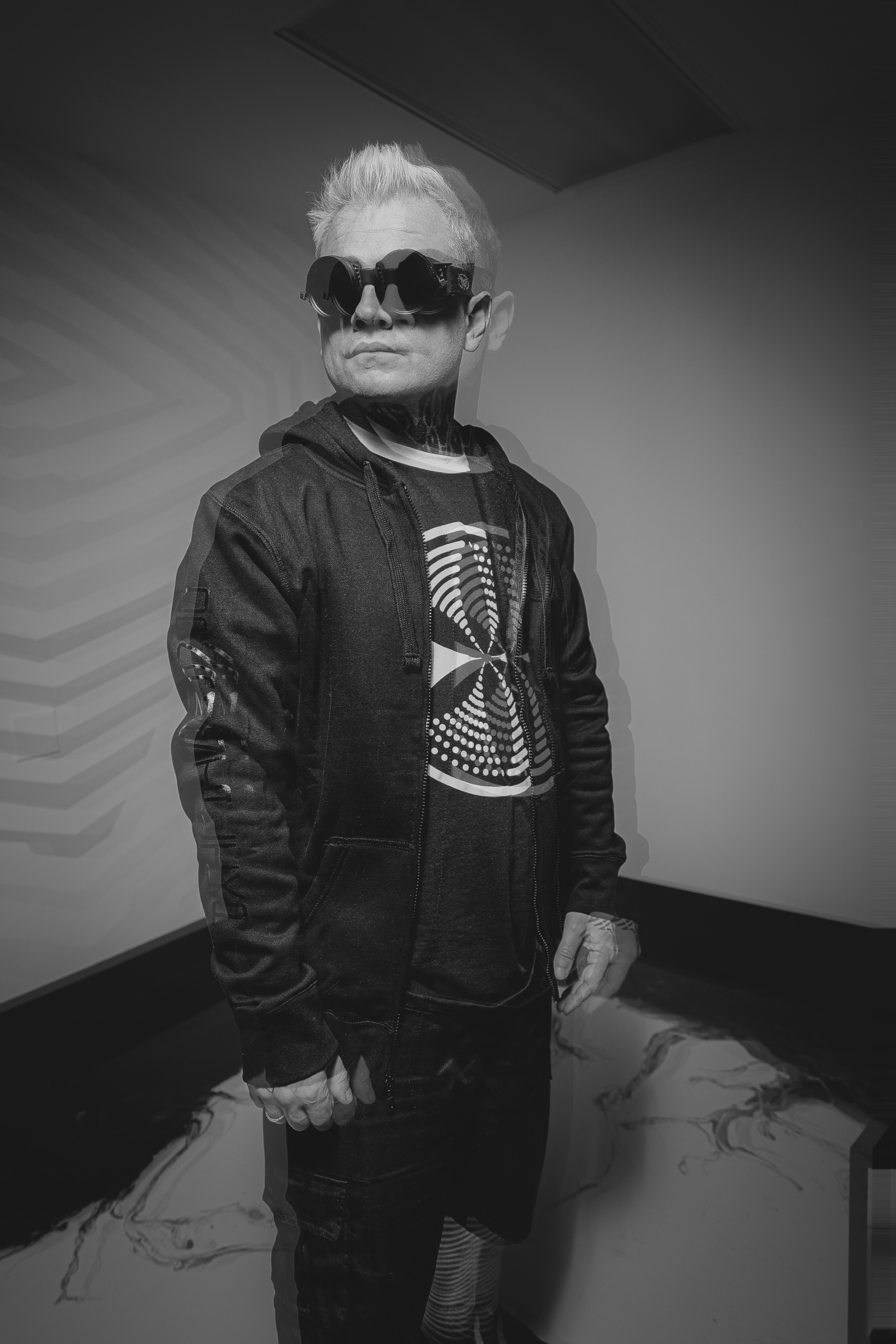 A person with short light-colored hair wearing sunglasses, a black jacket, a t-shirt with a tunnel-like design, and dark pants, standing against a plain wall.