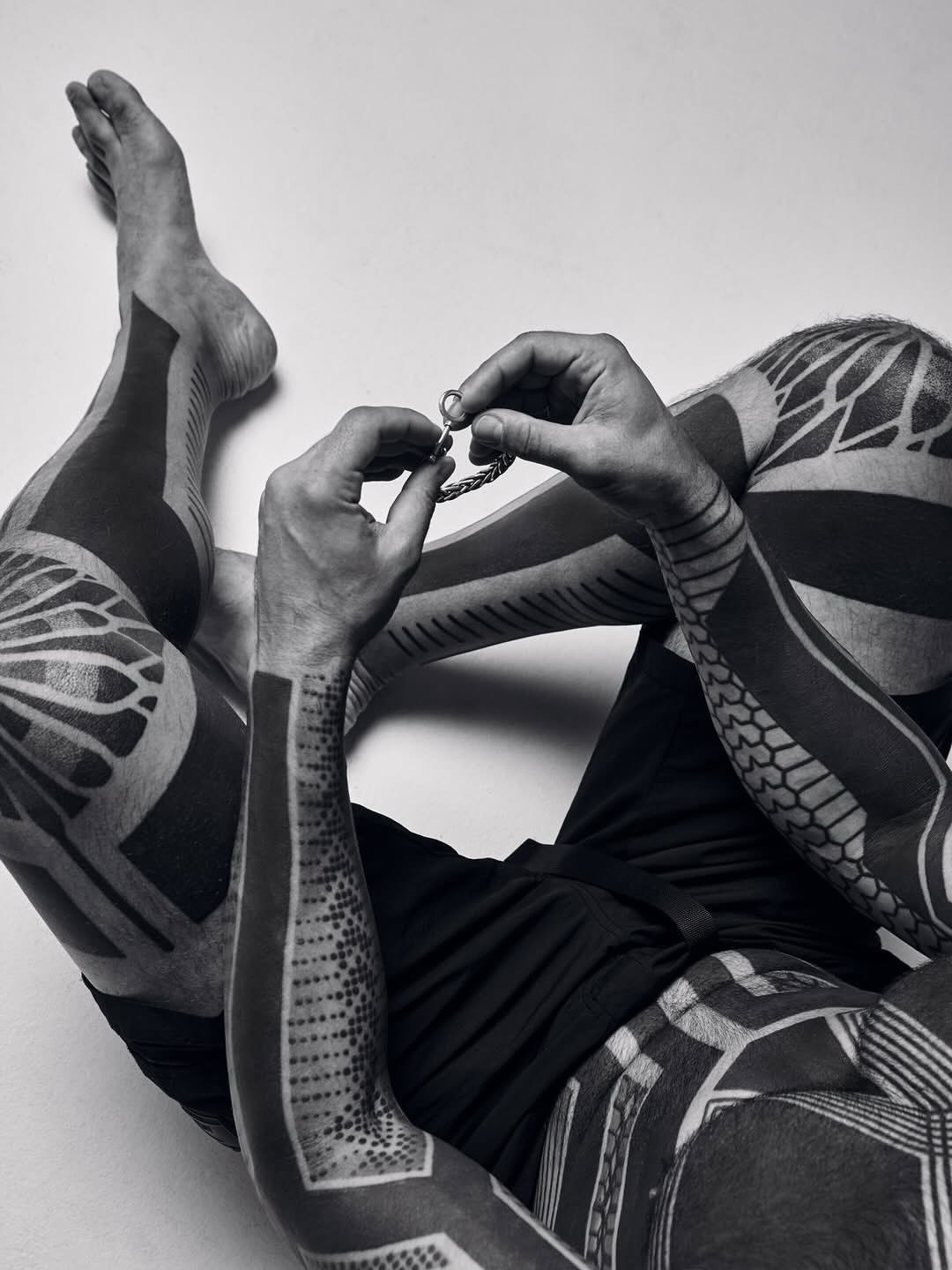 A person with tattoos on arms and legs, sitting on the floor, holding and examining a chain or bracelet.