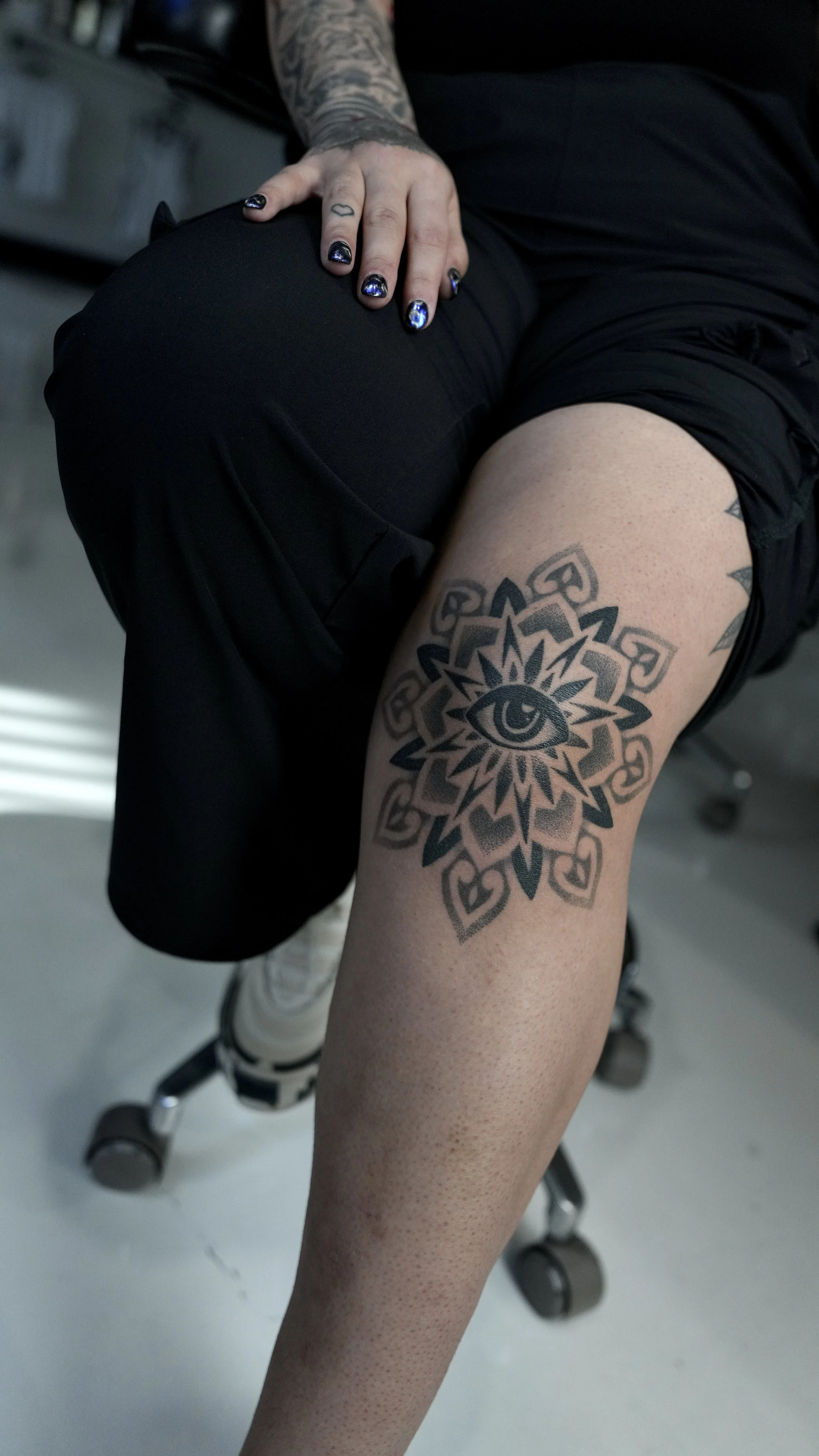 Close-up of a person's leg with a large tattoo of an eye surrounded by a geometric design and hearts, sitting on a chair with one hand resting on their knee.