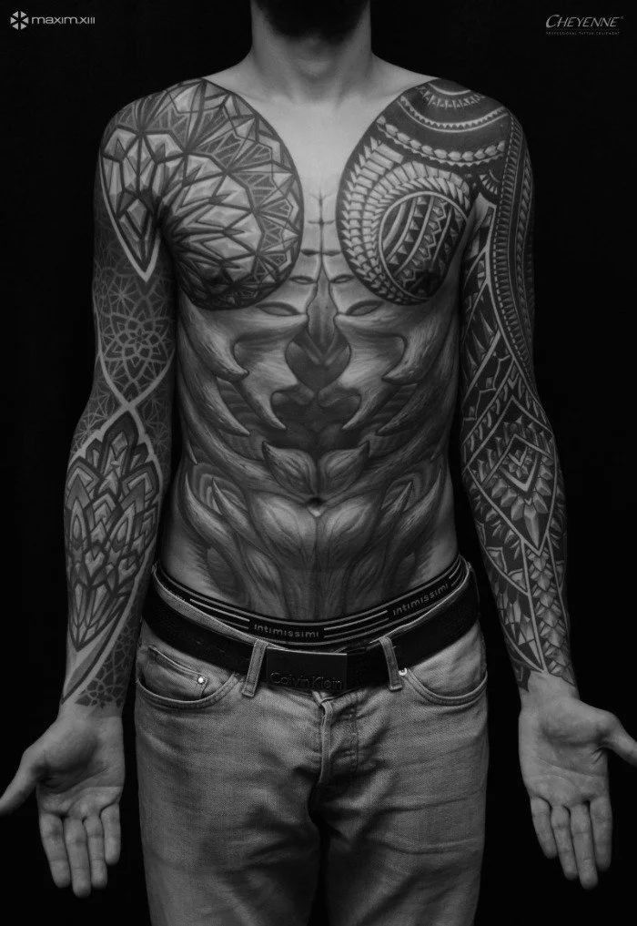 A person with extensive black and gray tattoos on their chest, stomach, and arms, wearing jeans and a Calvin Klein waistband, standing against a dark background.