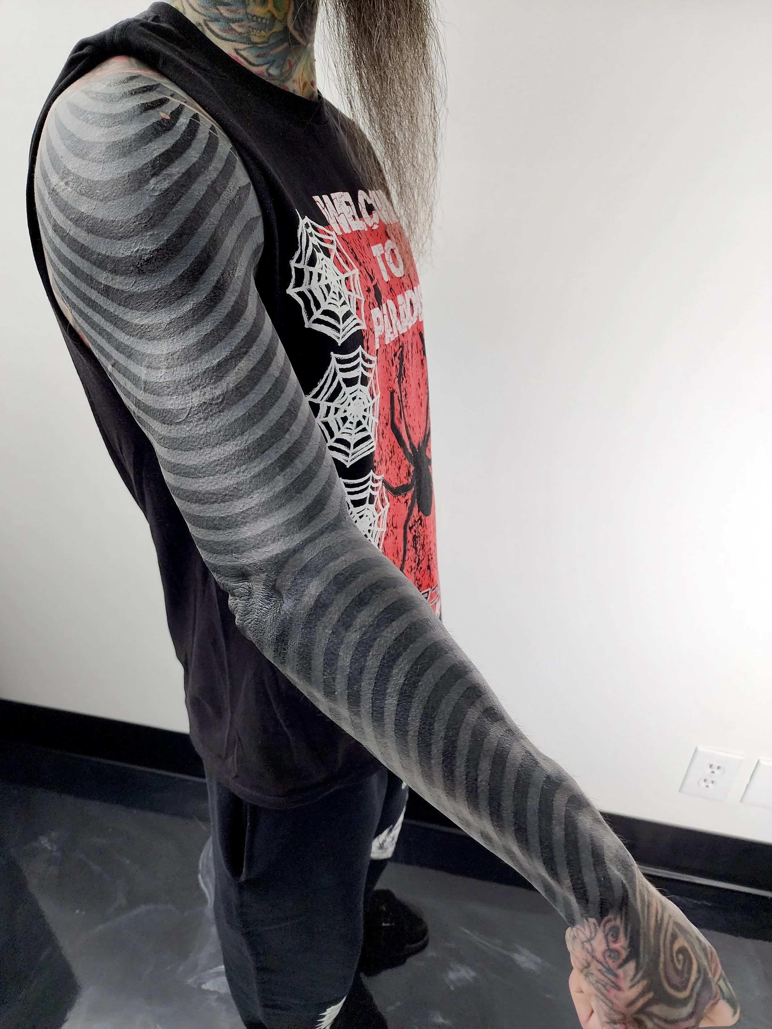 A person with extensive tattoos on their arm and neck wearing a sleeveless shirt with spider web designs on the sleeves and a graphic featuring a spider and text on the front.