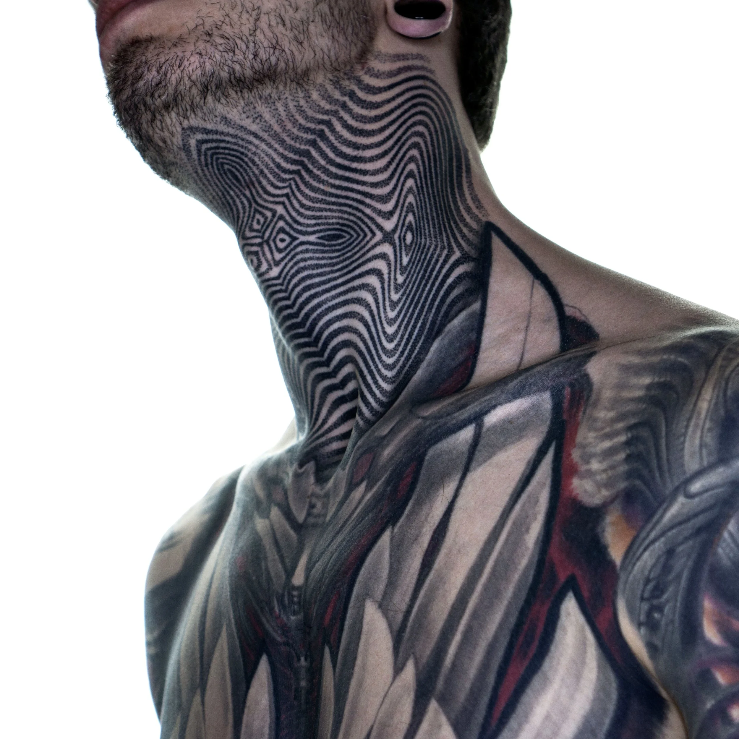 Close-up of a person's neck and upper chest with tattoos, including a black geometric pattern on the neck and colorful designs on the shoulder, and part of their beard and lips visible.