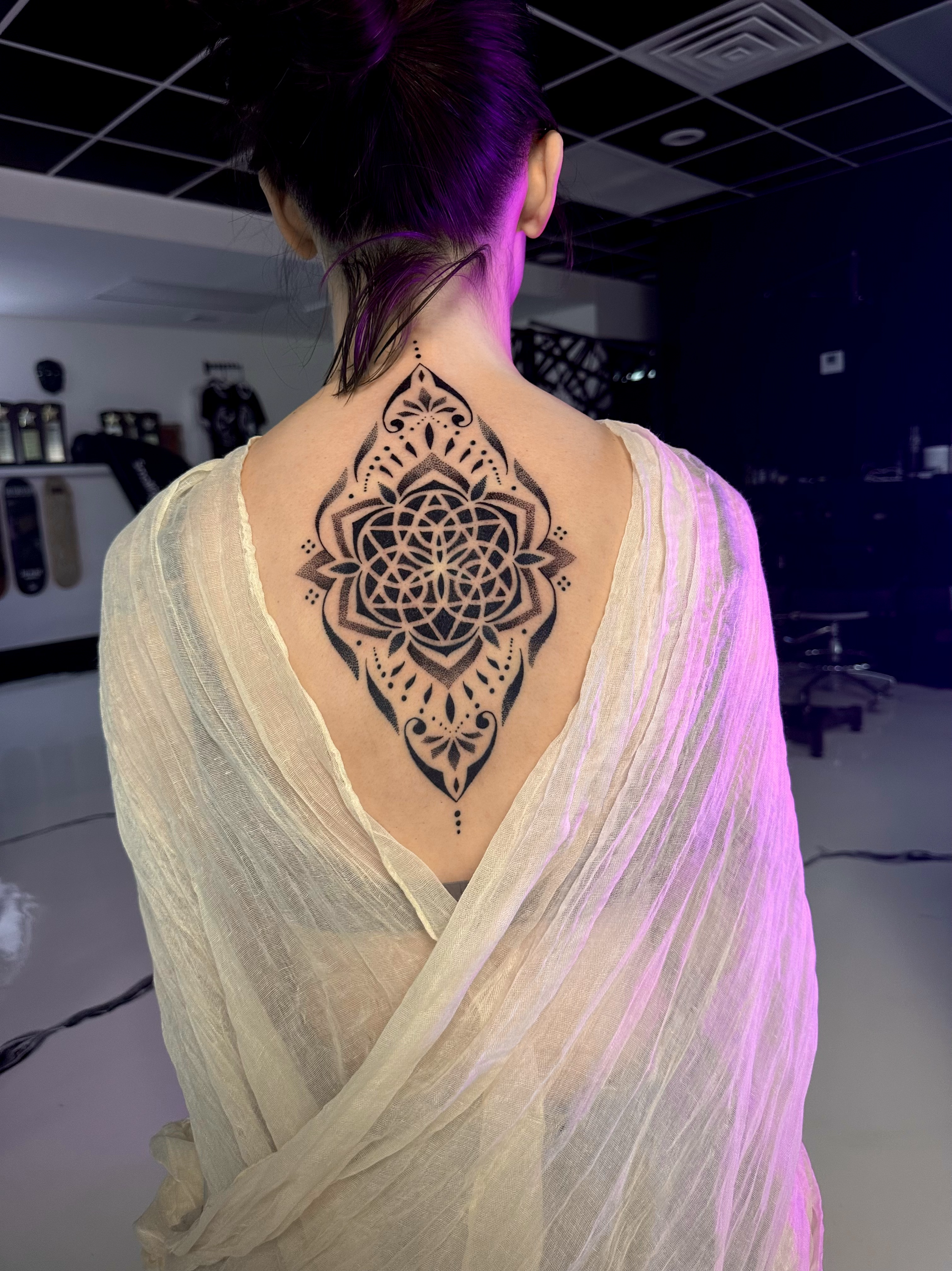 Back view of a woman with a large, intricate, black geometric and floral tattoo on her upper back, partially covered by a sheer, light-colored fabric.