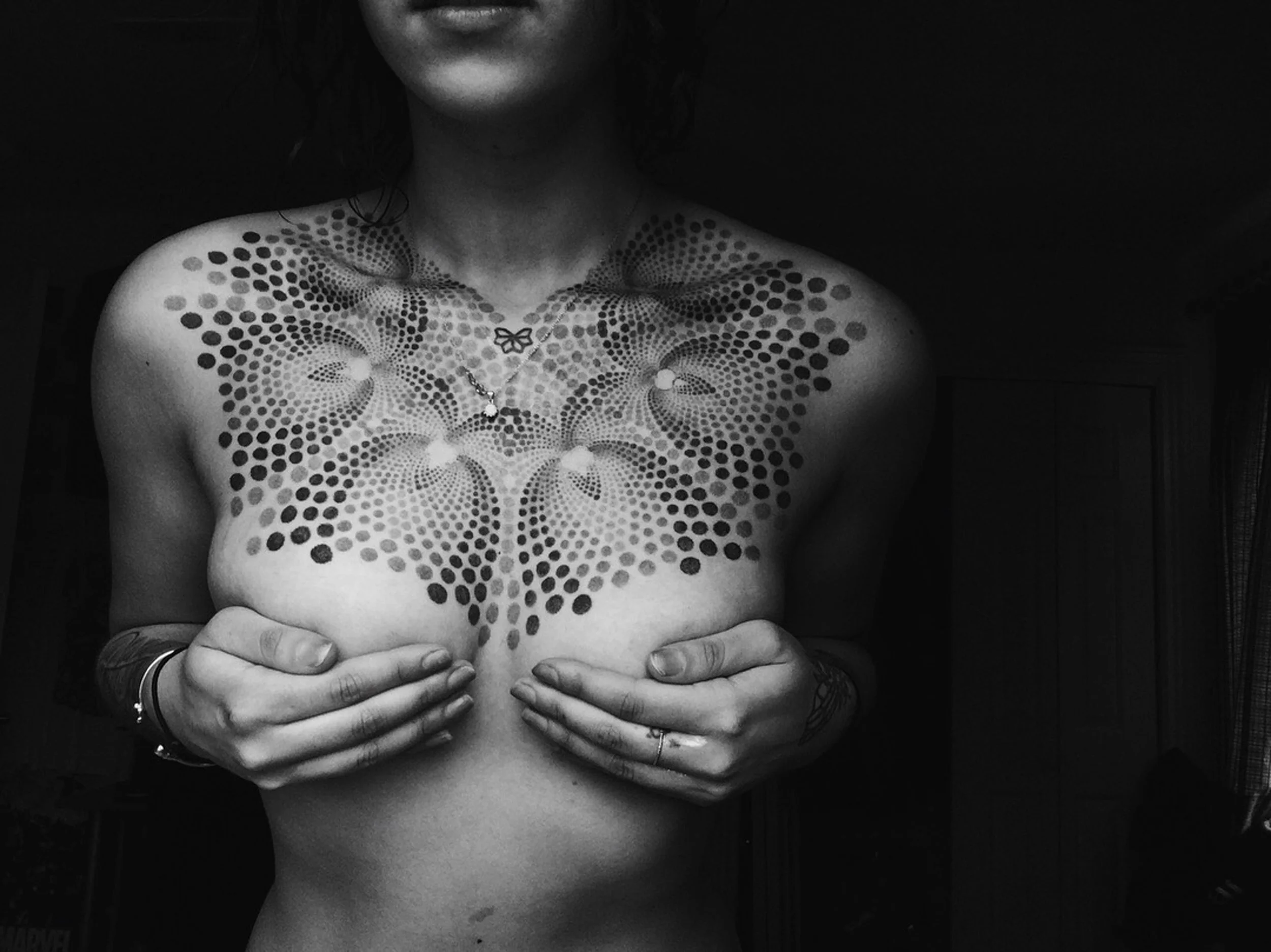 A person with a decorative tattoo on their chest covering their breasts, holding their breasts with hands. The tattoo features a butterfly and a pattern of dots creating a symmetrical design. The image is in black and white.