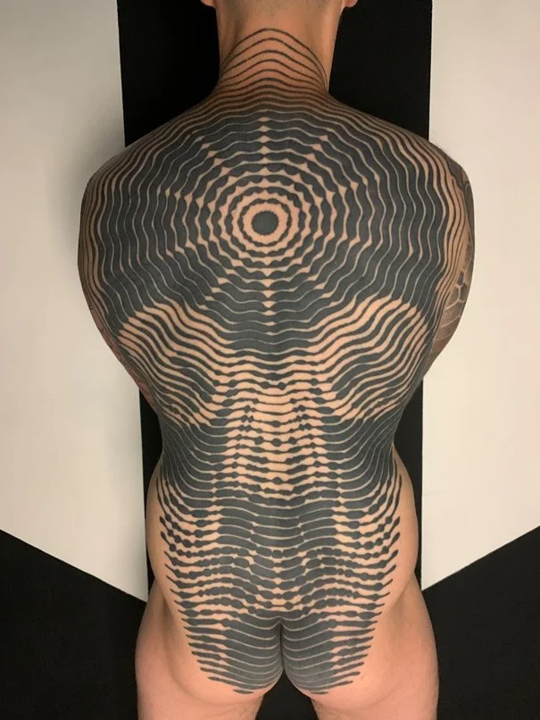 A person with a large, intricate black and beige geometric tattoo covering their back and shoulders, creating a hypnotic spiral pattern.