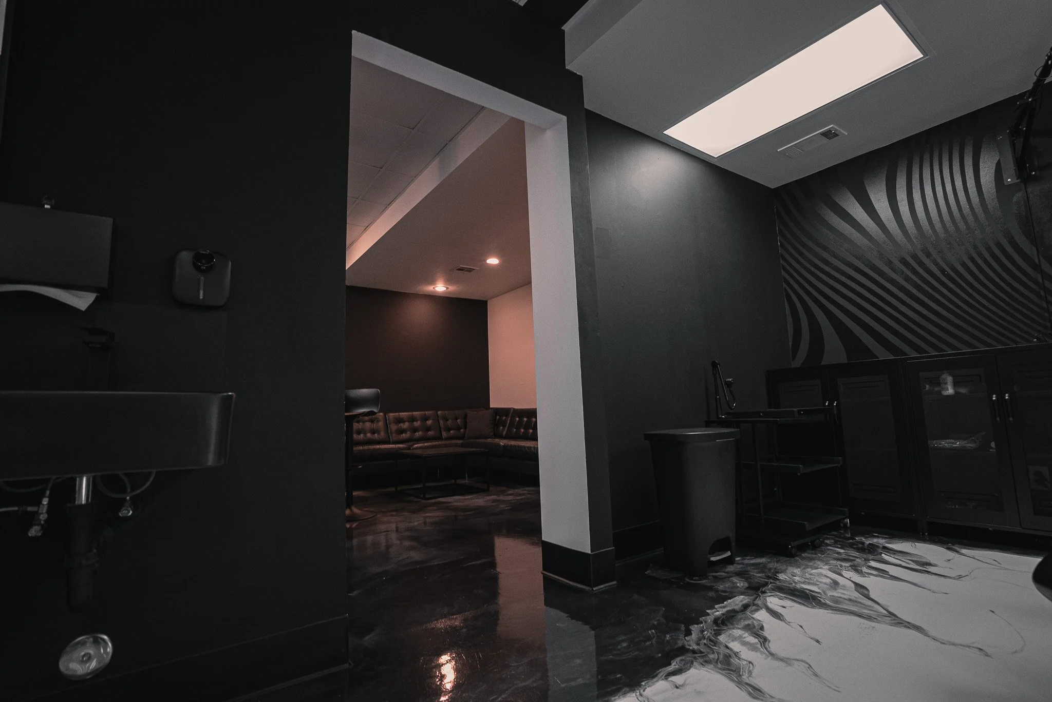 Interior view of a modern, dimly lit lounge or recreational room with black and gray decor, including leather seating, a black trash bin, a cabinet, and a small rolling cart.