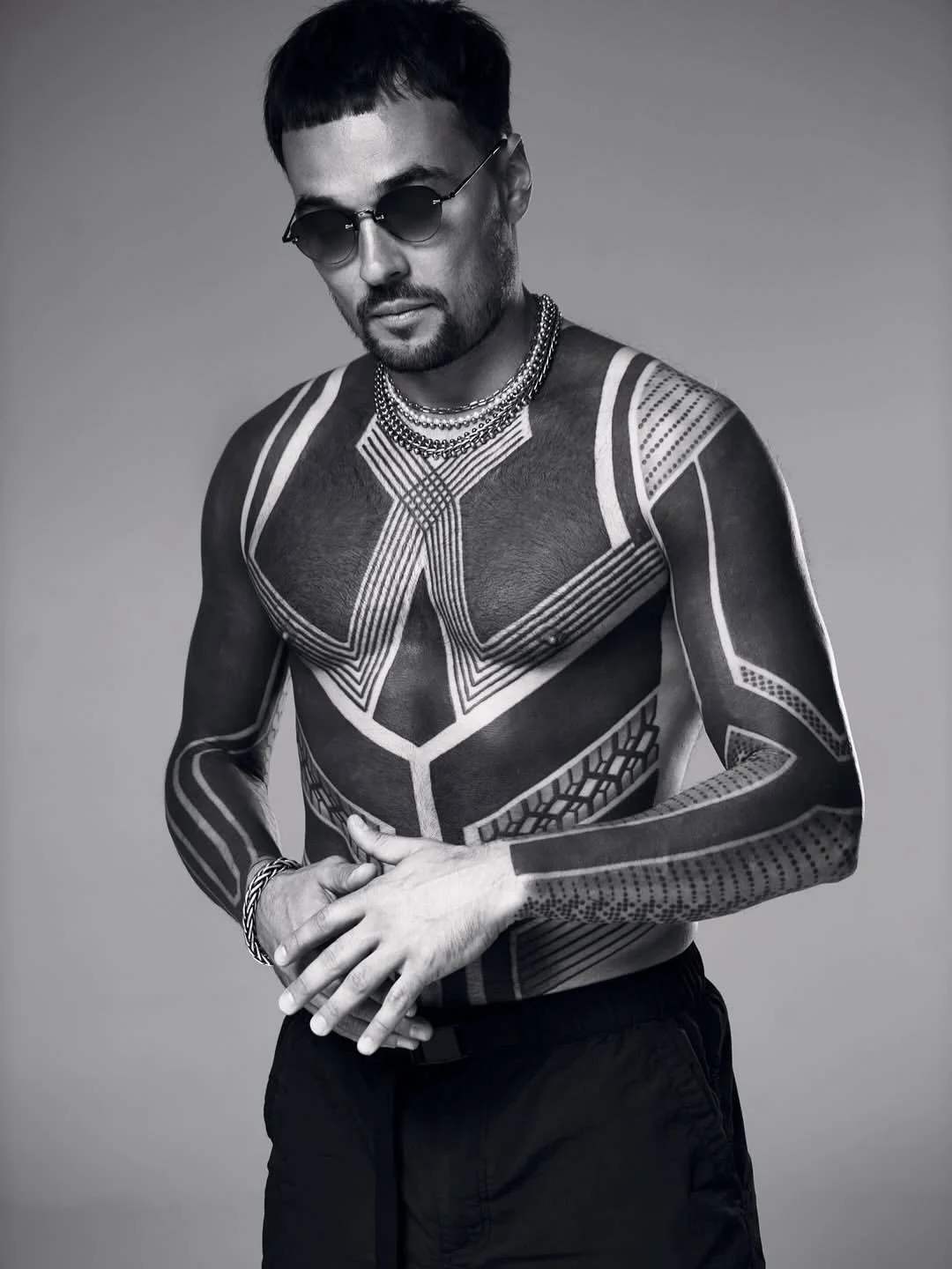 A man with tattoos covering his chest and arms, wearing sunglasses, jewelry, and dark pants, posing against a plain background in black and white.