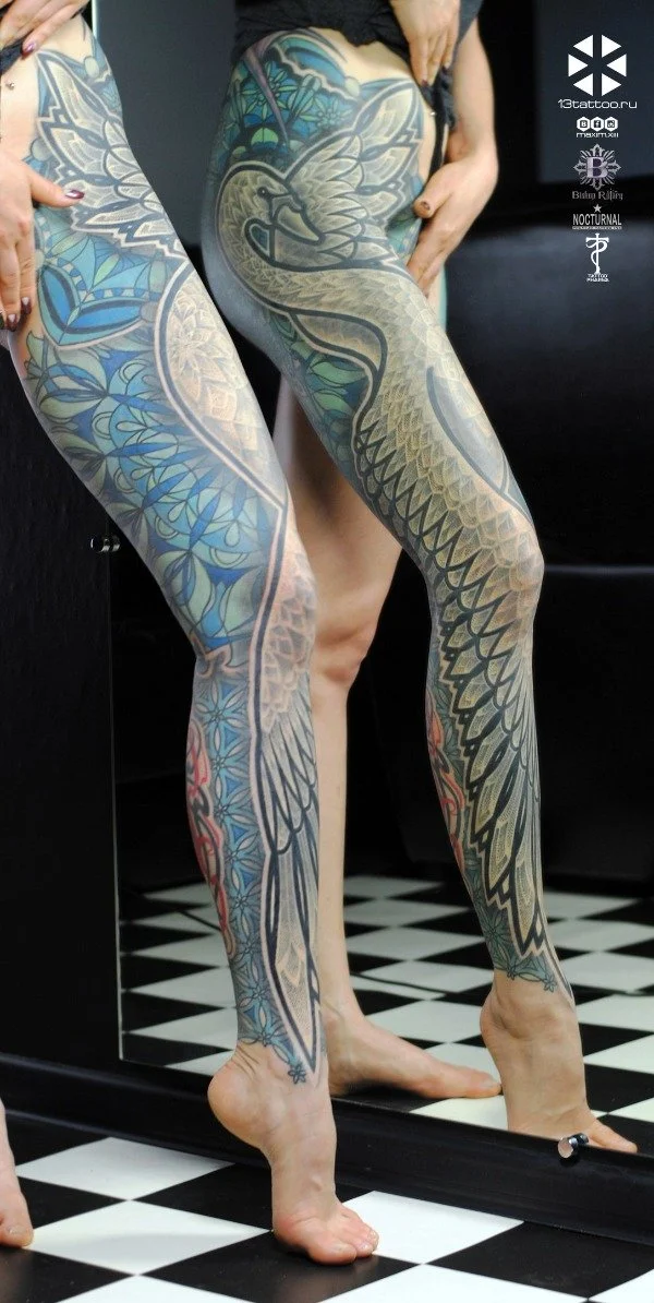 Close-up of two women with elaborate tattoos on their legs, including a swan and floral designs, standing on a black-and-white checkered floor, with a mirror reflecting their legs.