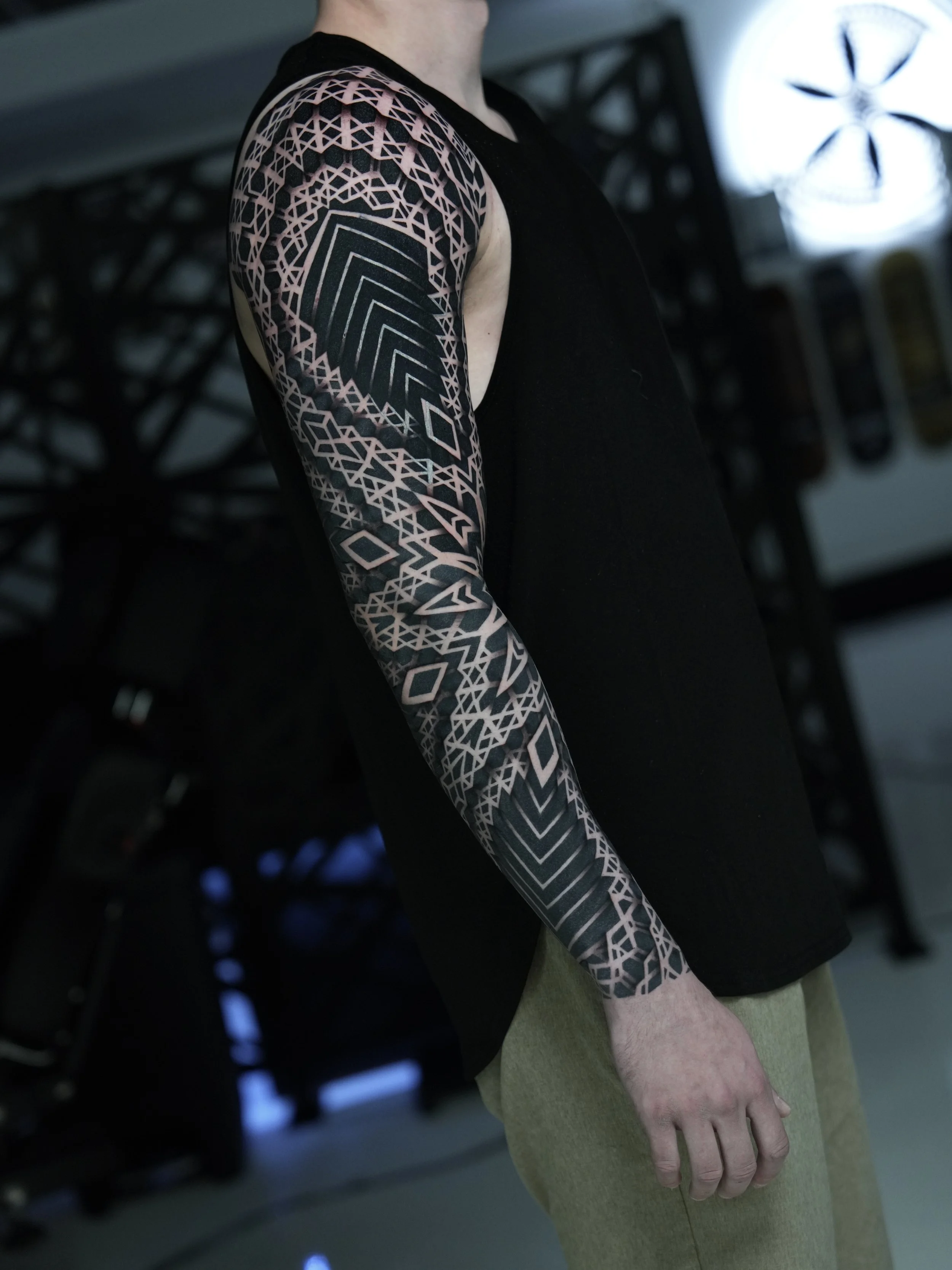 A person with a geometric tattoo sleeve on their arm, wearing a black sleeveless shirt and beige pants, standing indoors with a black lattice structure in the background.