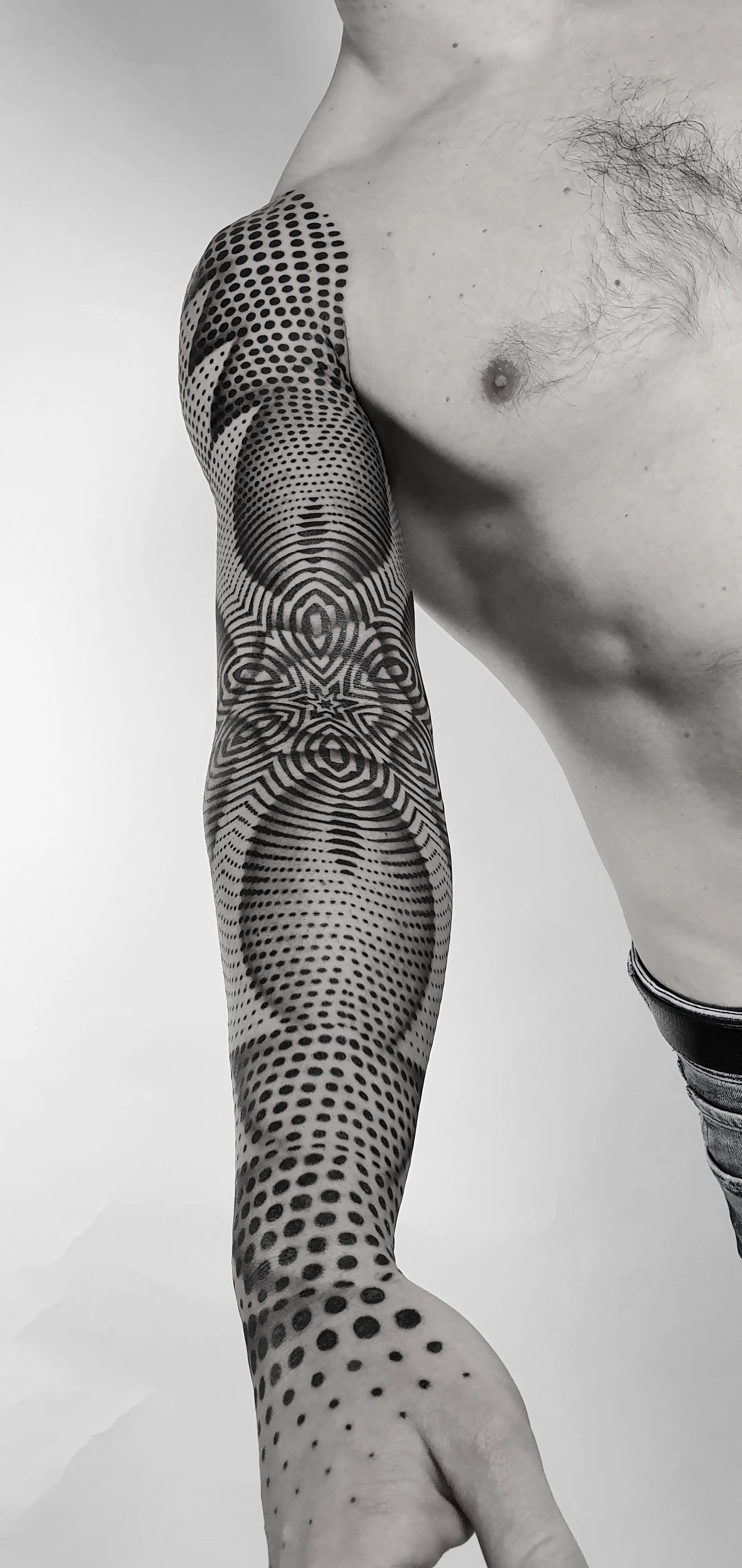 A shirtless person showing a detailed black and gray geometric and dot tattoo on their arm. The tattoo extends from the shoulder to the wrist.