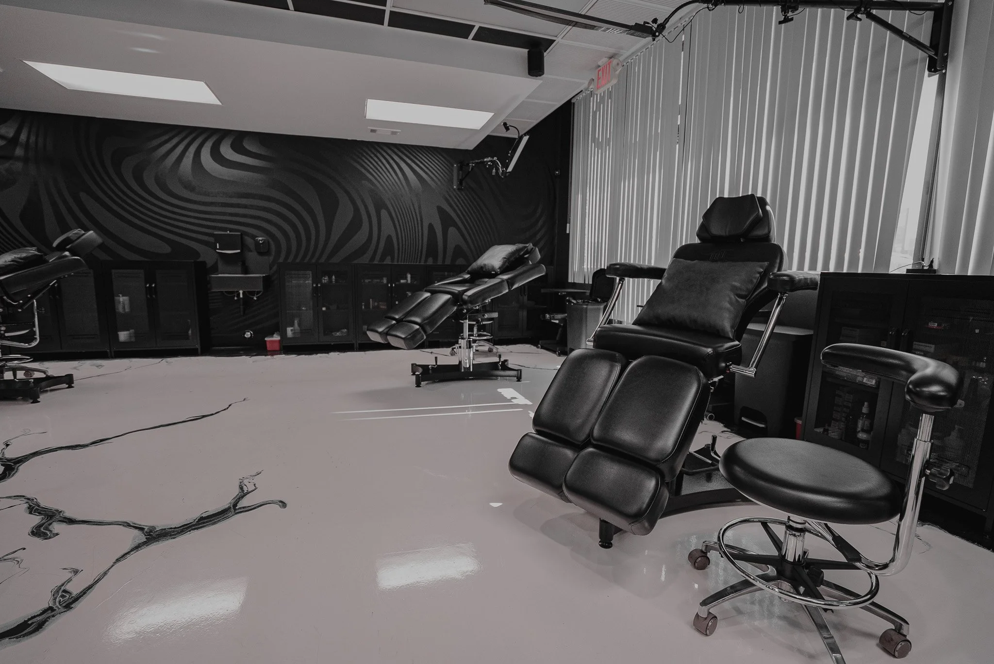 Medical treatment room with black reclining chairs, cabinets, and a patterned black and white wall, illuminated by ceiling lights and natural light from window blinds.