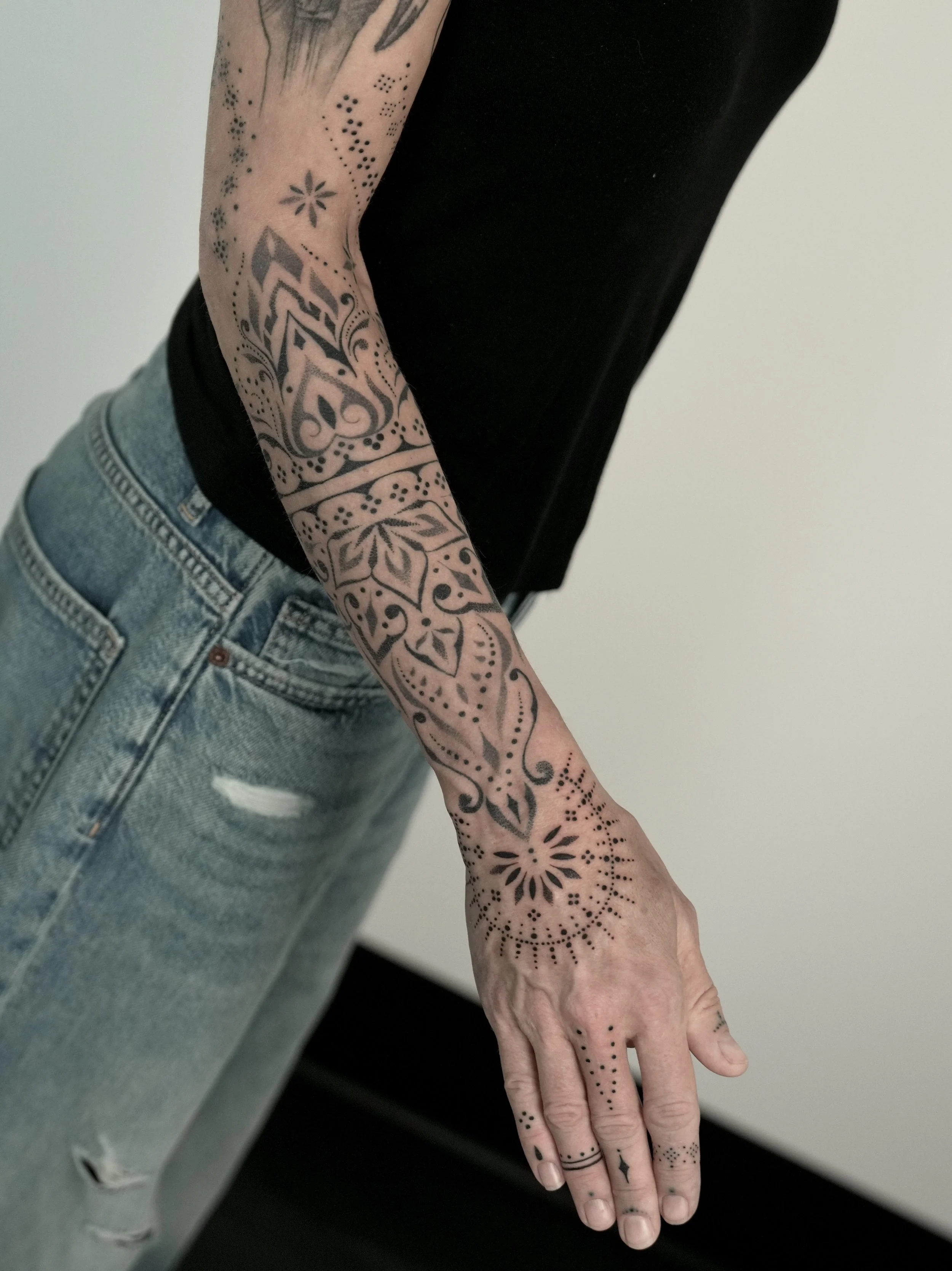 A person with a detailed black tattoo sleeve on their arm and hand, wearing a black shirt and light blue ripped jeans, standing against a plain white wall.