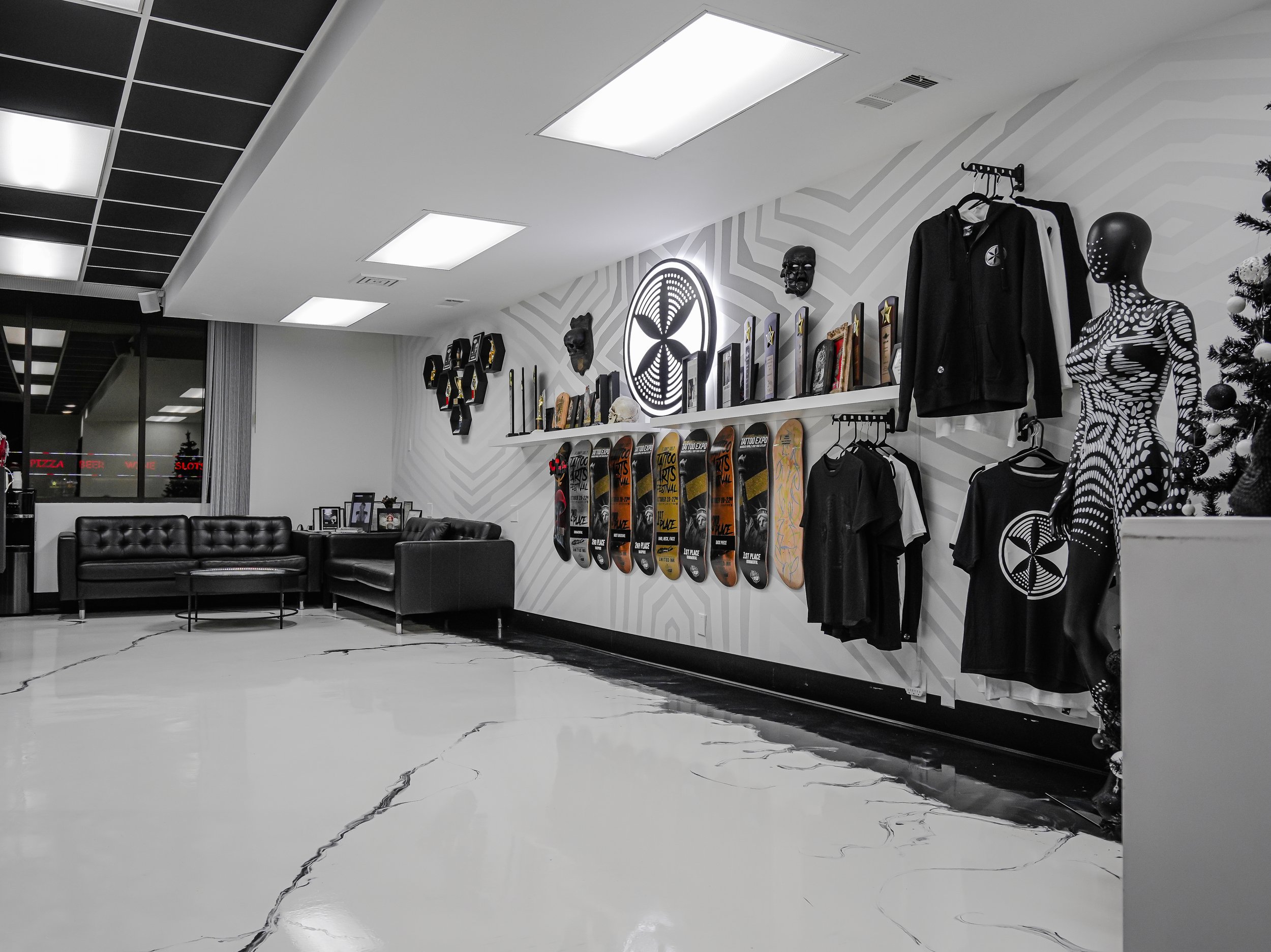 Indoor lounge area with black leather couches, a coffee table, and framed photos, adjacent to a retail display wall featuring skateboards, clothing, masks, sculptures, and a decorated Christmas tree.
