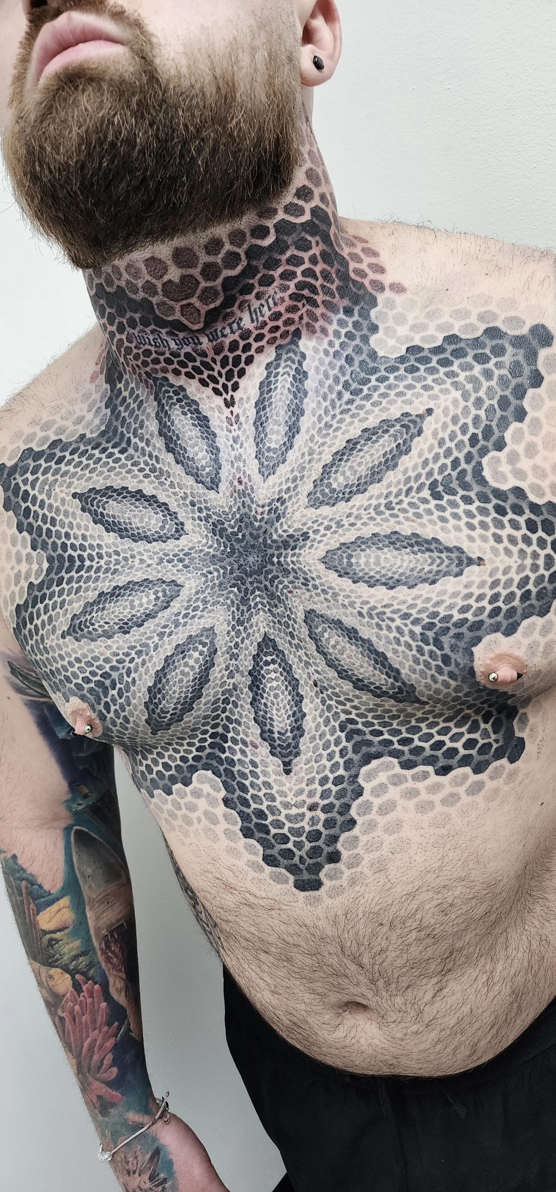 A shirtless man with a large chest tattoo featuring a detailed, symmetrical geometric pattern in black and gray. He has a beard, a piercing on his ear, and an arm tattoo with colorful floral and landscape designs.