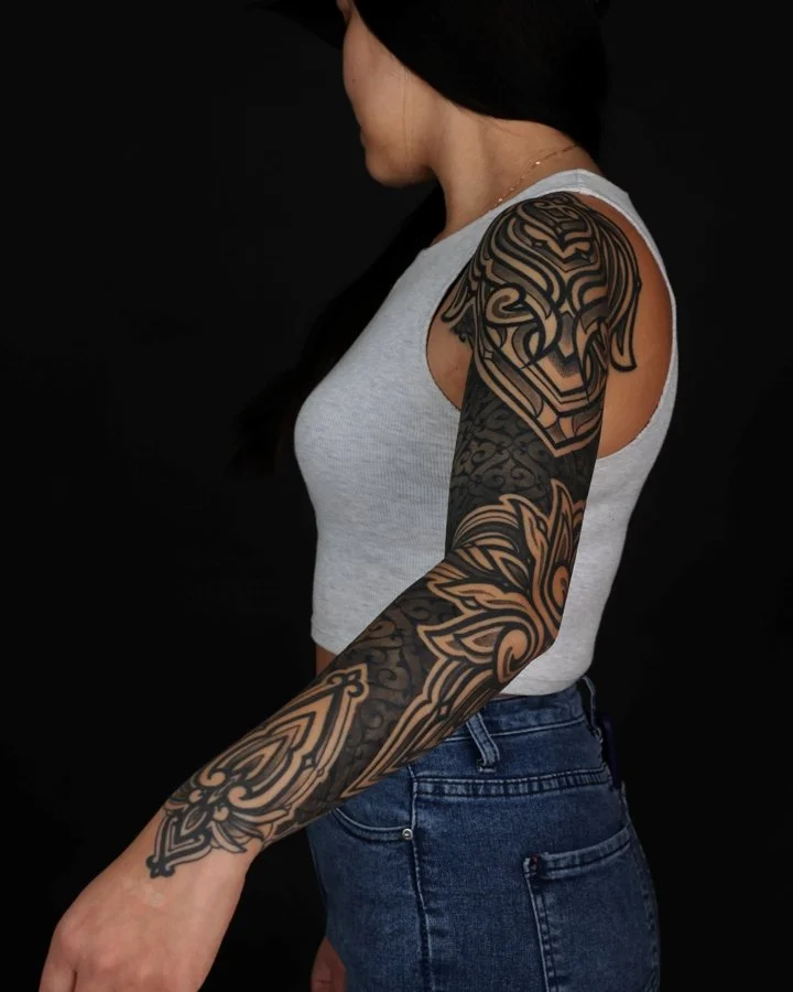 A woman with long dark hair wearing a gray sleeveless top and blue jeans, showing her detailed black and tan tattoos on her arm against a black background.