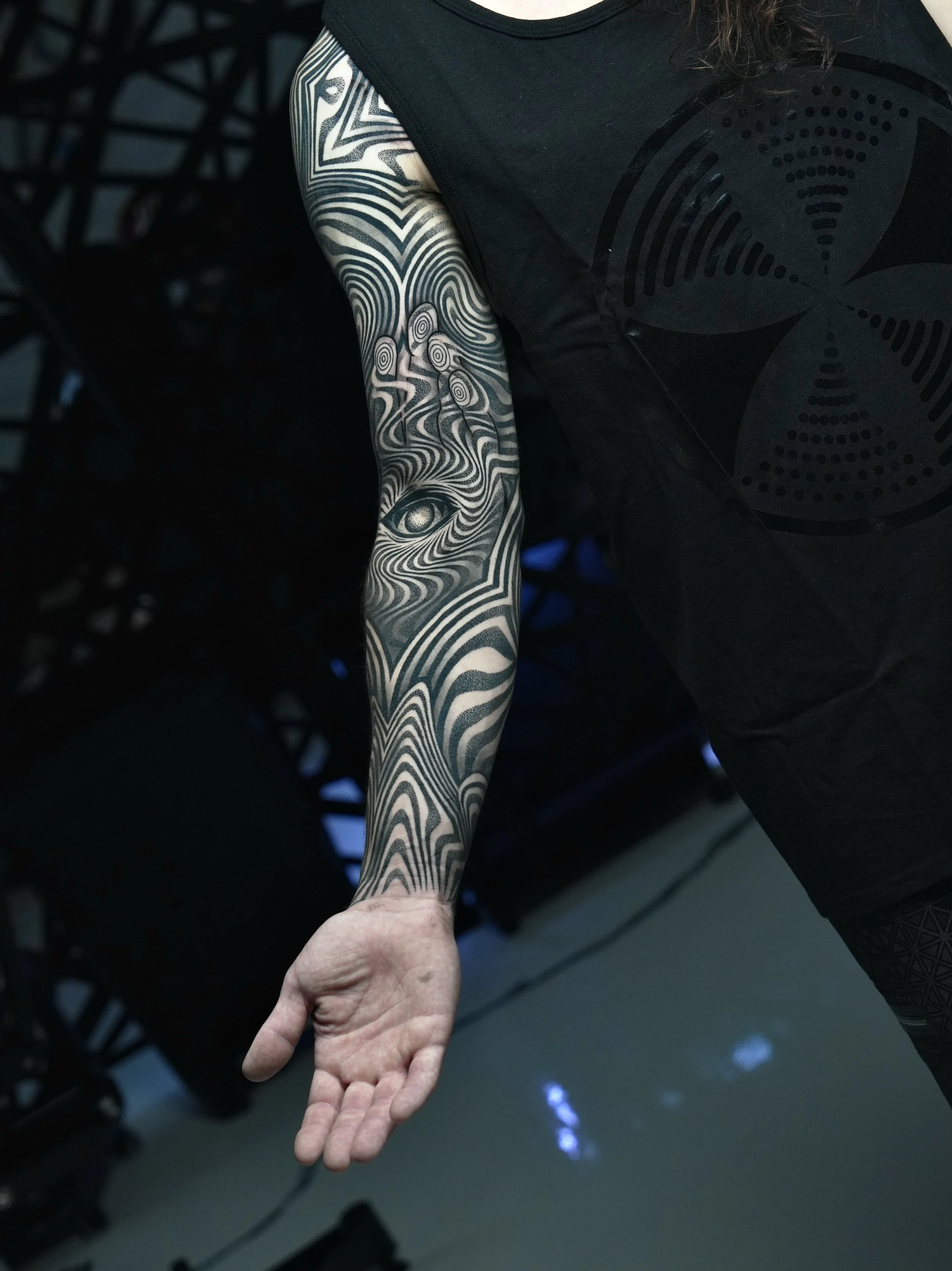 A person with a black tattoo sleeve featuring intricate line patterns and an eye design on their arm, wearing a black sleeveless shirt.
