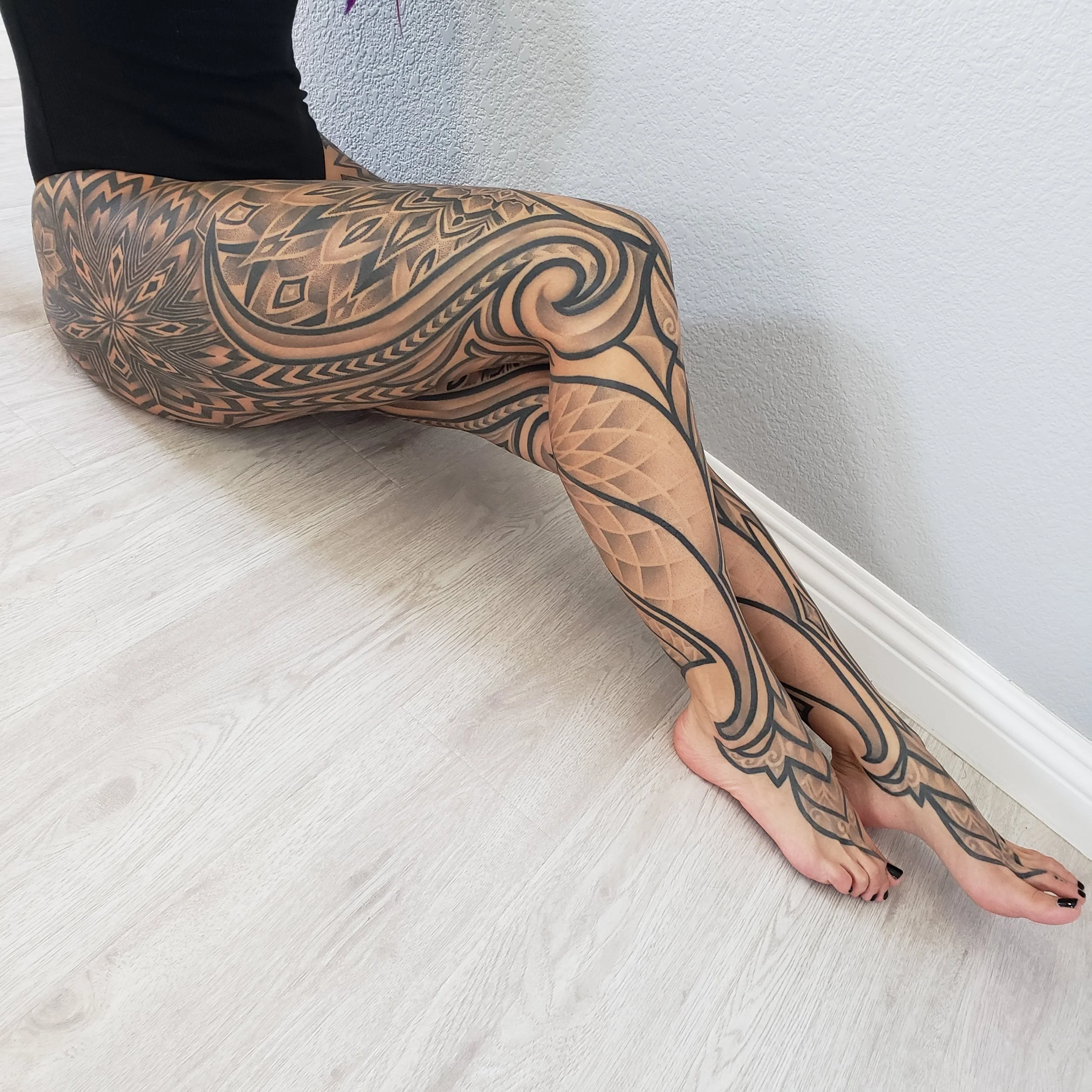 A person with extensive black tattoo art on their legs, sitting on a light-colored wooden floor, with one leg extended and bent at the knee, wearing black nail polish.