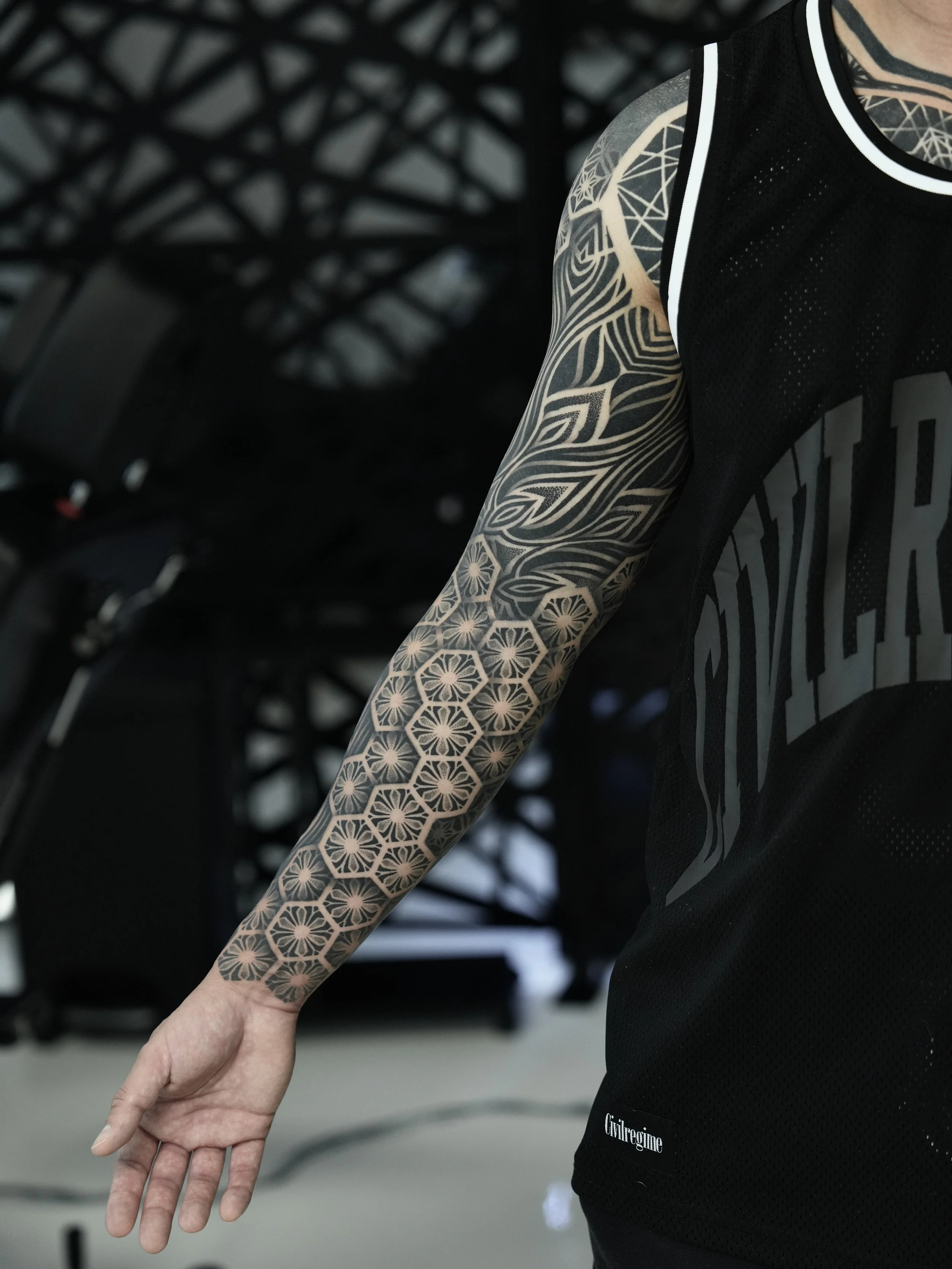 Close-up of a person's arm with a black geometric and floral tattoo sleeve, wearing a black sleeveless sports jersey.