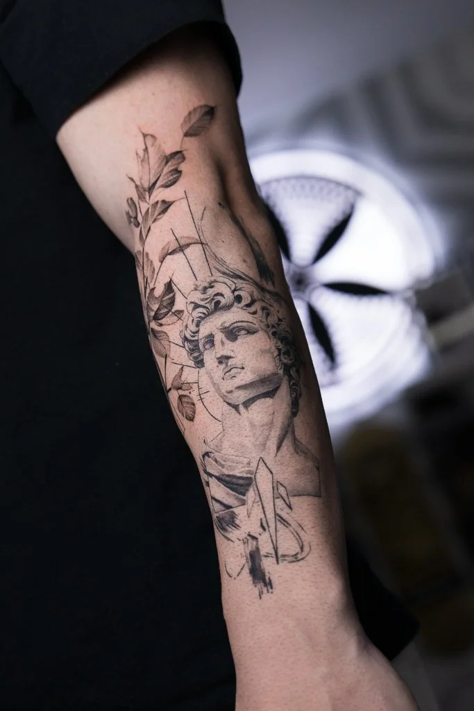 Tattoo of a classical-style sculpture of a woman's head on a person's forearm, surrounded by leaves and abstract lines.