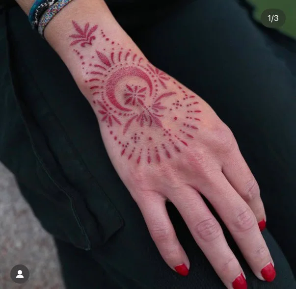 Close-up of a hand with a red henna tattoo design on the back, featuring floral and circular patterns.