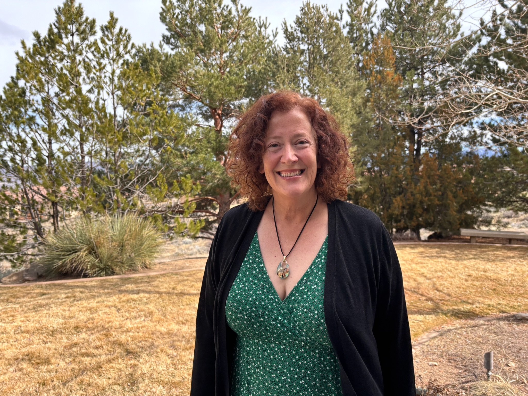 Dr Wendy Lafever Jordanov smiling outdoors in a park, wearing a green dress with white polka dots, a black cardigan, and a pendant necklace, with trees and grass in the background.
