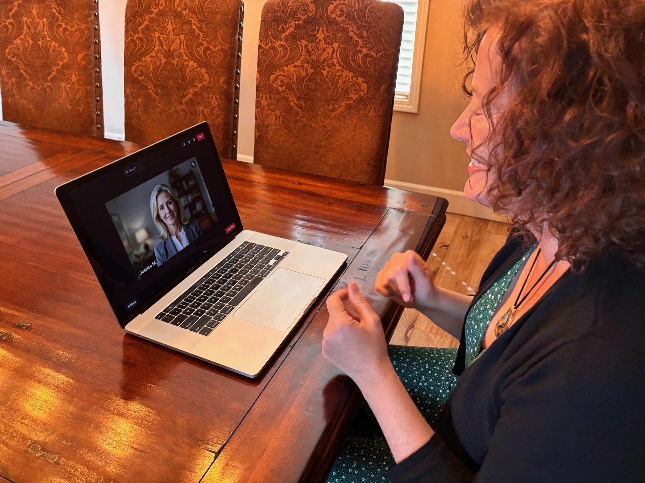 Dr Wendy Jordanov sits at a wooden table during a video call on her laptop, showing a smiling woman with shoulder-length blonde hair on the screen.