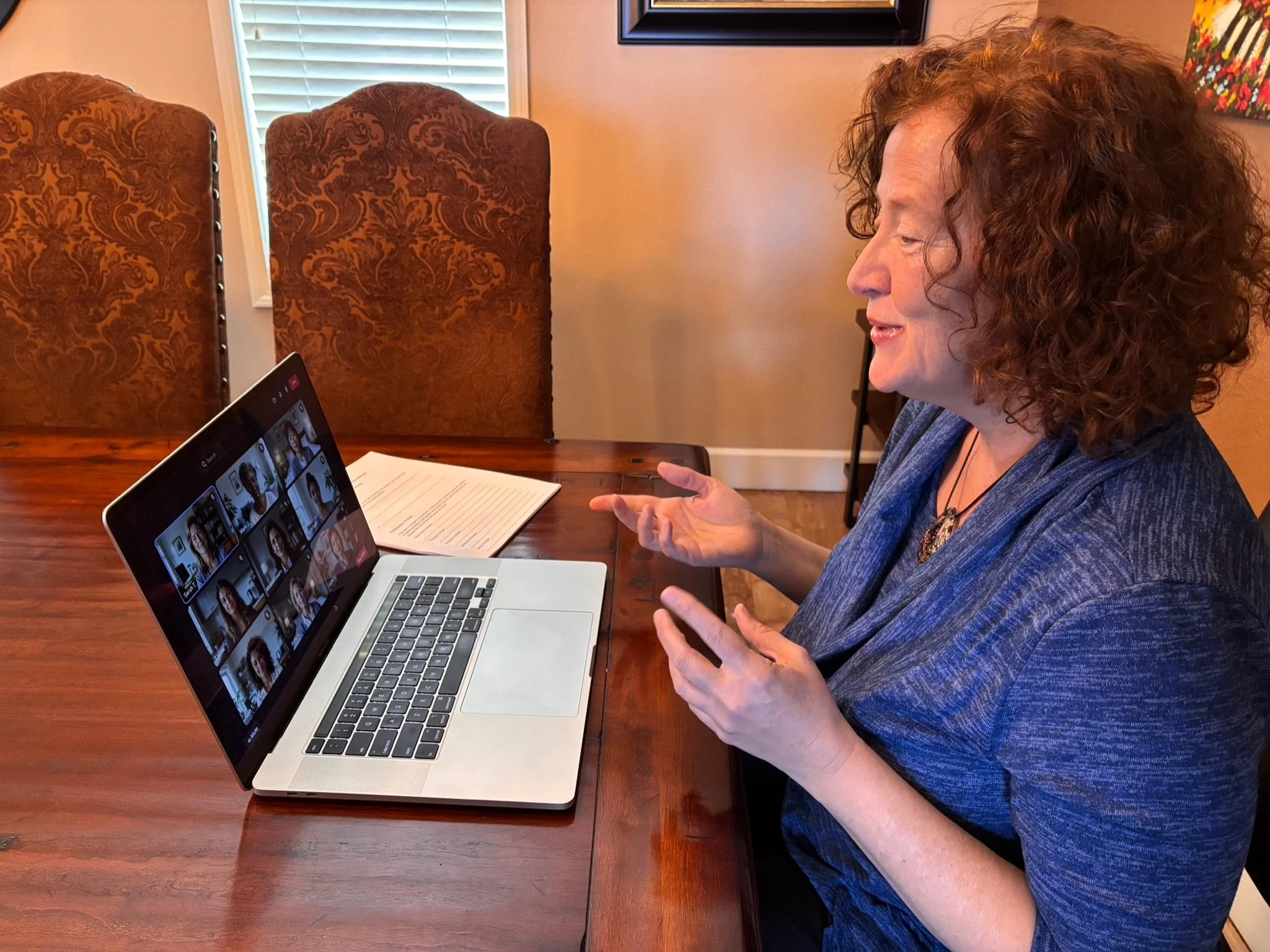 Dr Wendy Lafever Jordanov sits at a wooden table on a video conference call on her laptop. She is gesturing with her hands and appears engaged. Behind her are orange patterned chairs and a window with blinds.