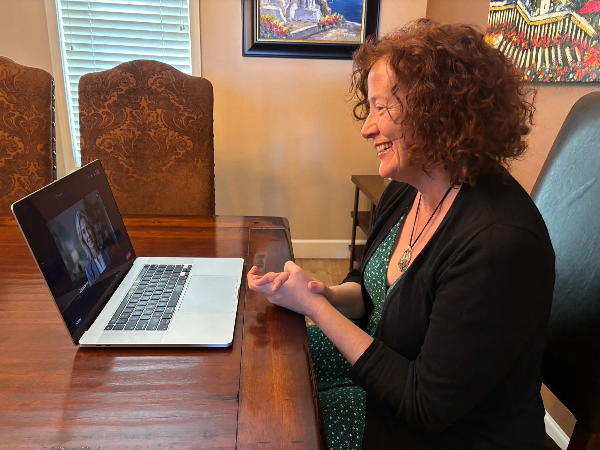 Dr Wendy Jordanov smiling and talking to someone on a video call on her laptop, seated at a wooden table in a room with framed artwork on the walls.