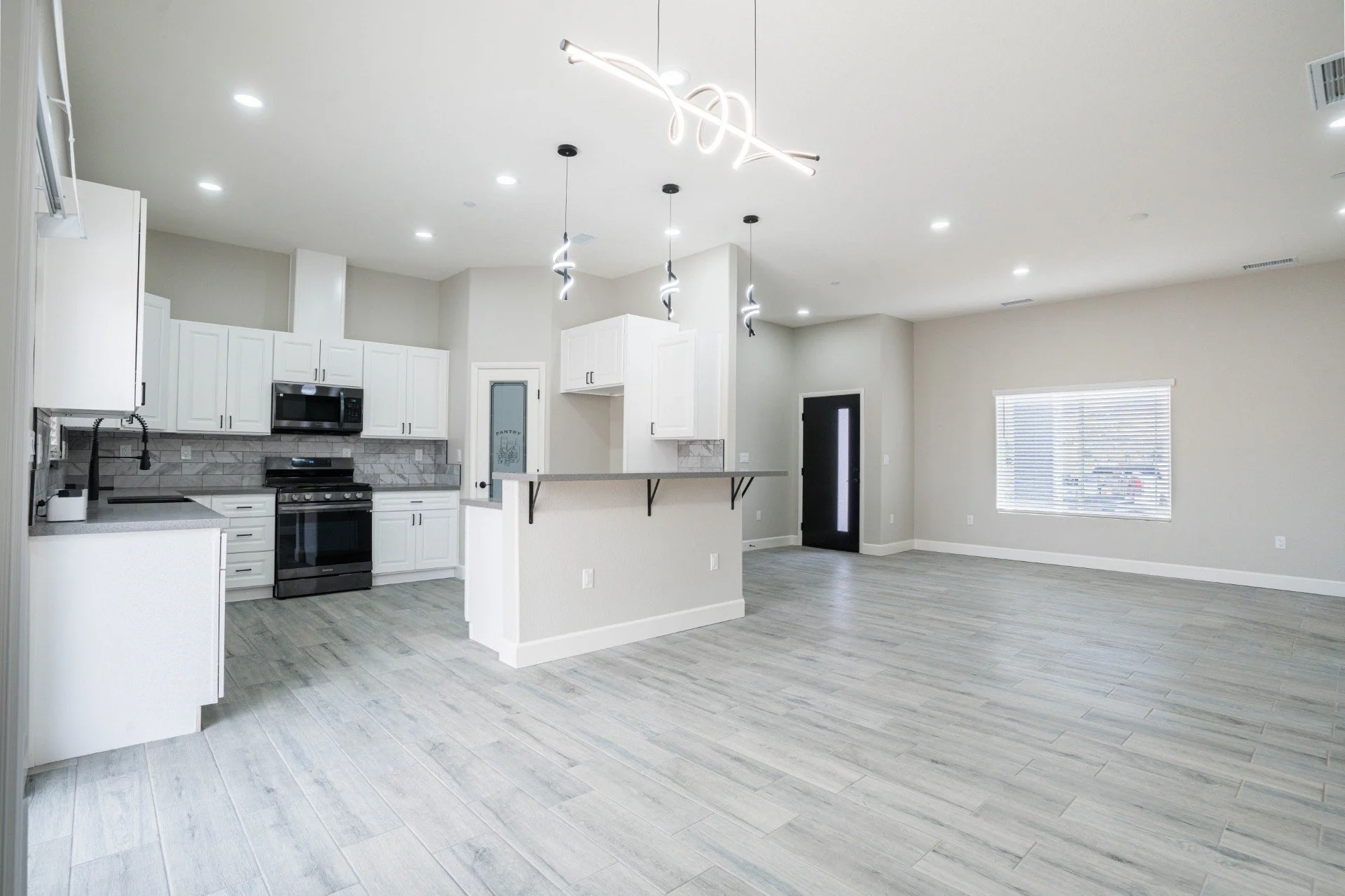 Open-concept kitchen with white cabinets, stainless steel appliances, and light wood flooring. Modern light fixtures hang from the ceiling, and there is a kitchen island with a breakfast bar. A window with blinds lets in natural light, and a black do