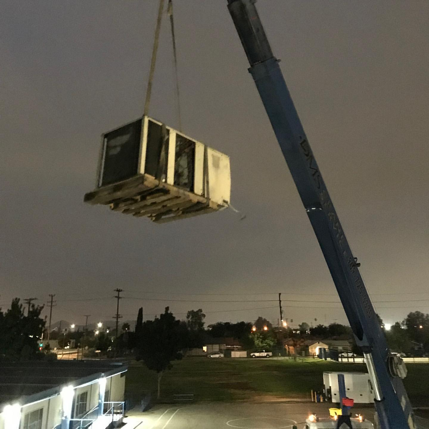 A crane lifting an air conditioning unit at night above a parking lot, with residential houses and trees in the background.
