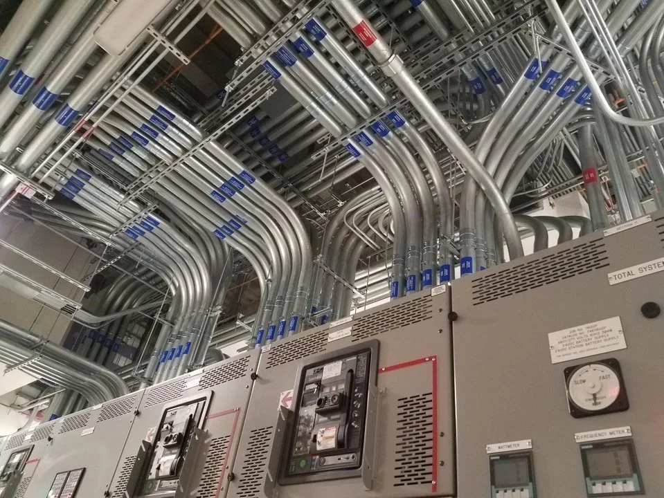 Electrical control panels with numerous silver conduits and wiring running overhead in an industrial setting.