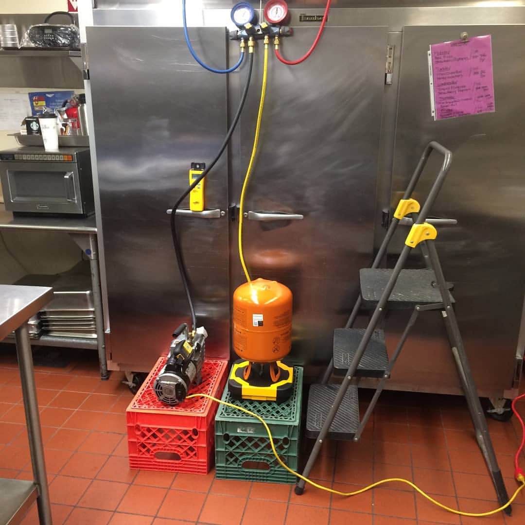 Propane tank connected to a yellow heating device, with a pressure meter and hoses, near a large stainless steel refrigerator, in a commercial kitchen.