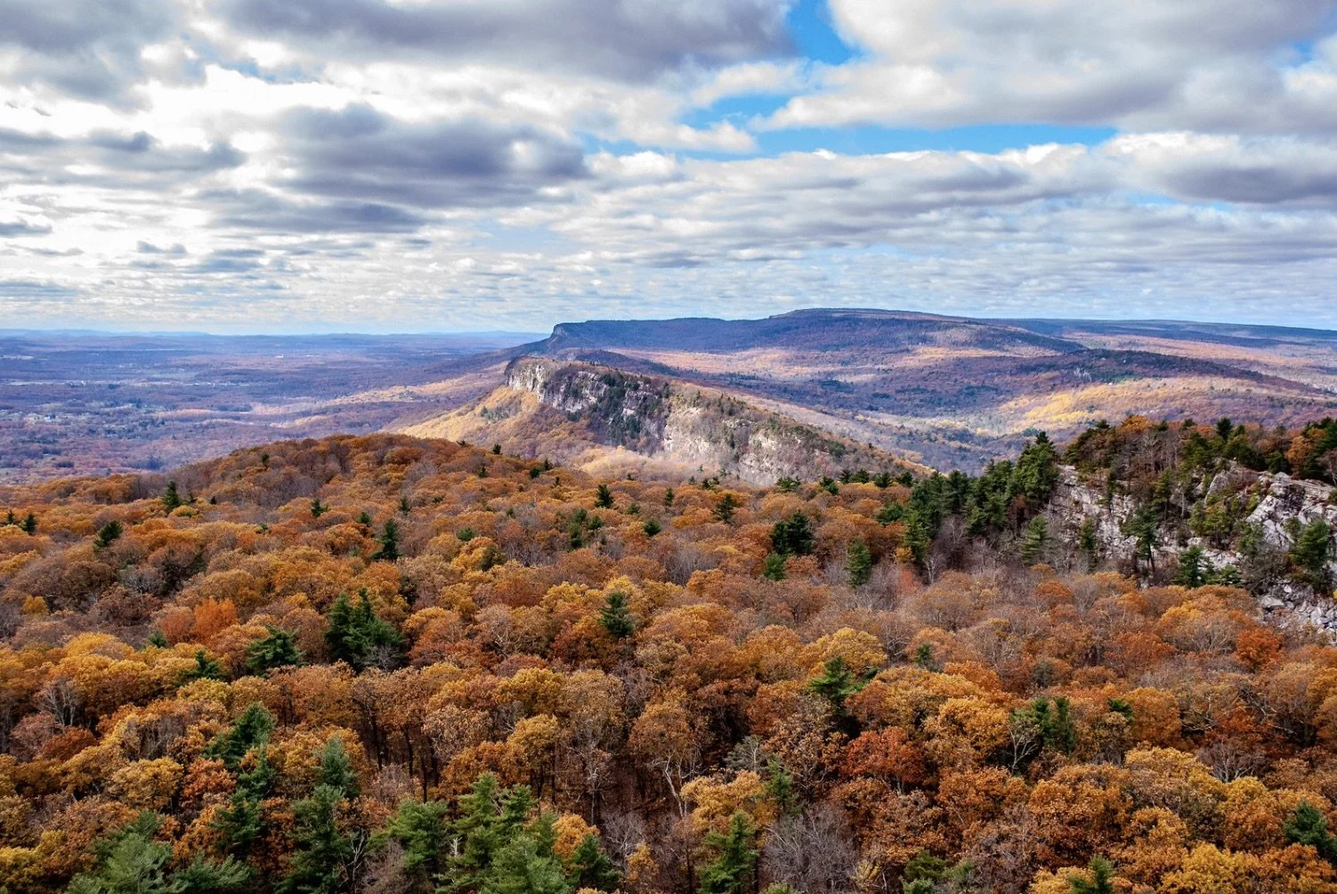 Scenic view of a mountainous landscape with orange, yellow, and green fall foliage, rocky cliffs, and a partly cloudy sky.
